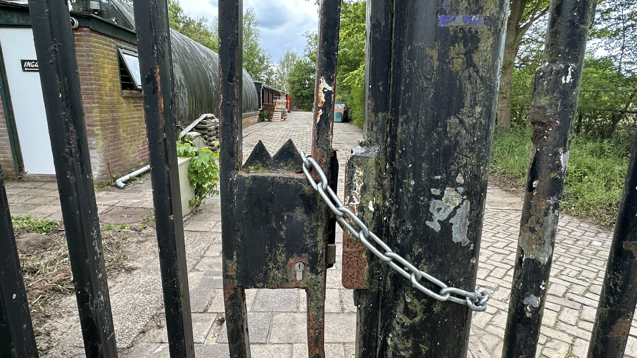 Verroest hek met ketting en slot, zicht op gebouw met bordje "Ingang" en een verhard pad tussen bomen.