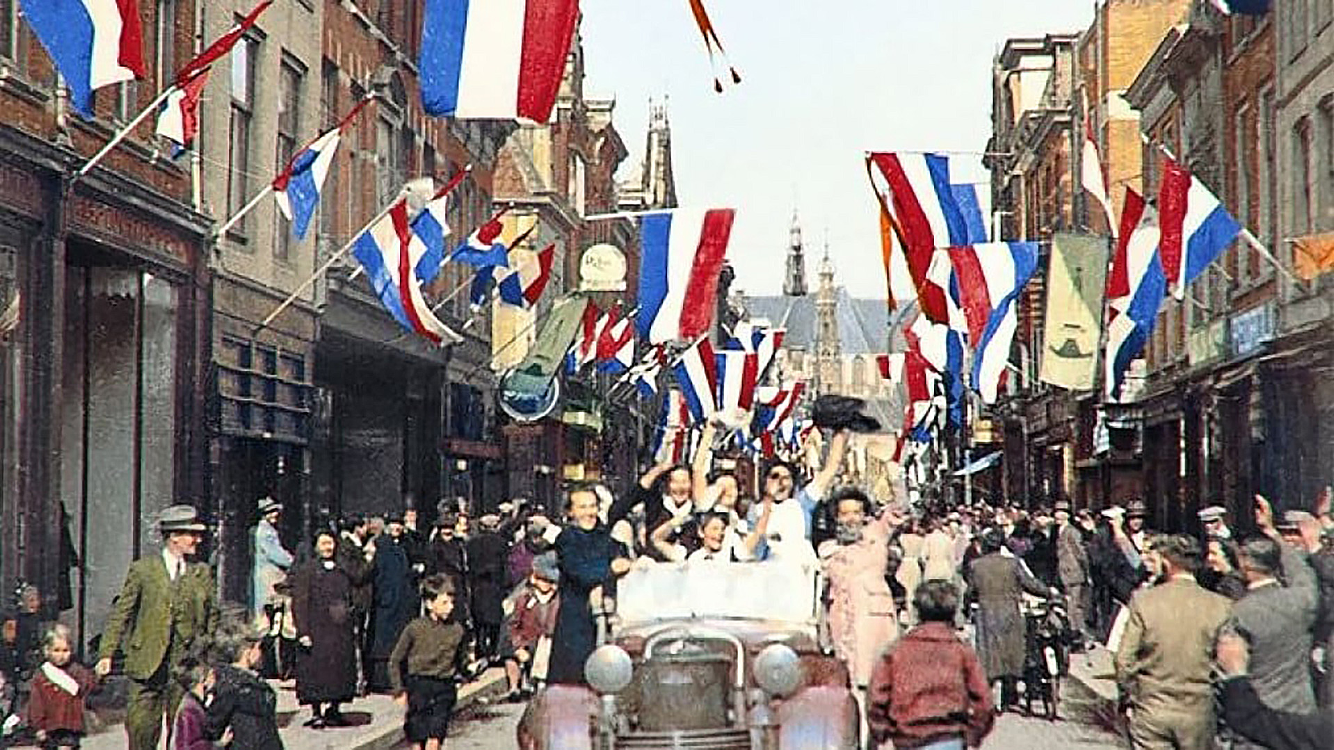 Drukke straat vol Nederlandse vlaggen en feestvierende mensen, met een auto vol mensen die juichen en zwaaien.