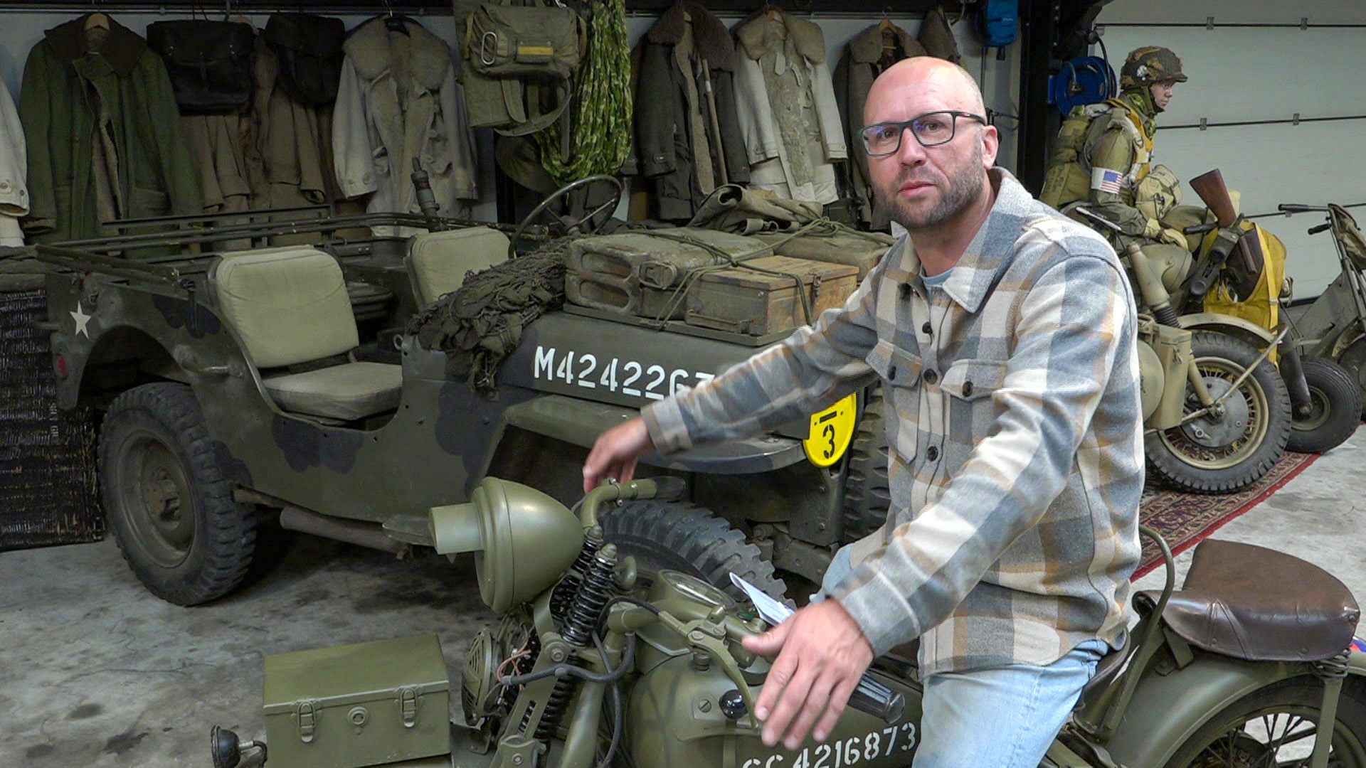 Man zit op een vintage militaire motorfiets naast een oude militaire jeep, met uniformen en uitrusting op de achtergrond.