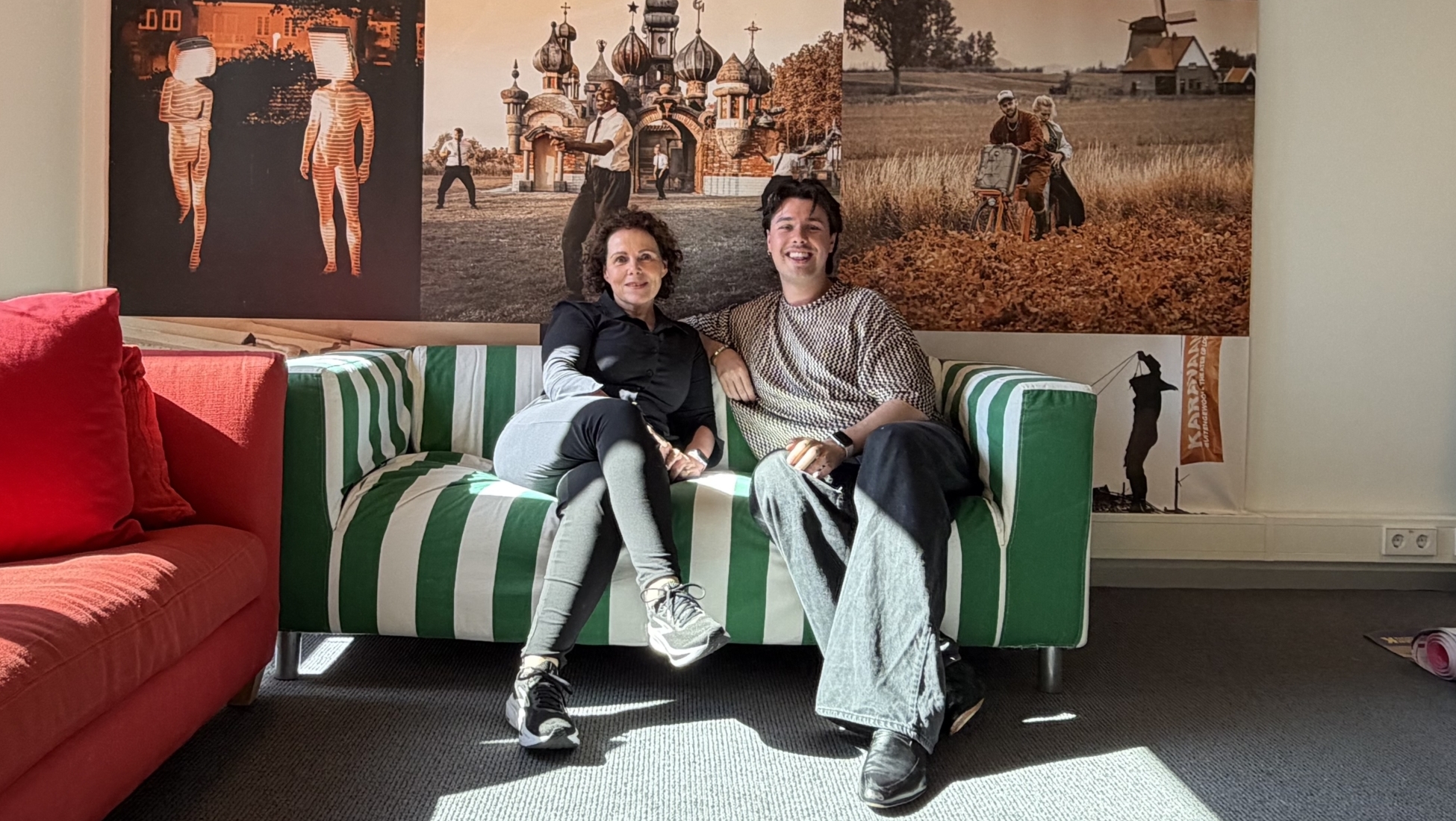 Twee mensen zitten op een groen-wit gestreepte bank in een kantoor met een grote fotocollage aan de muur.