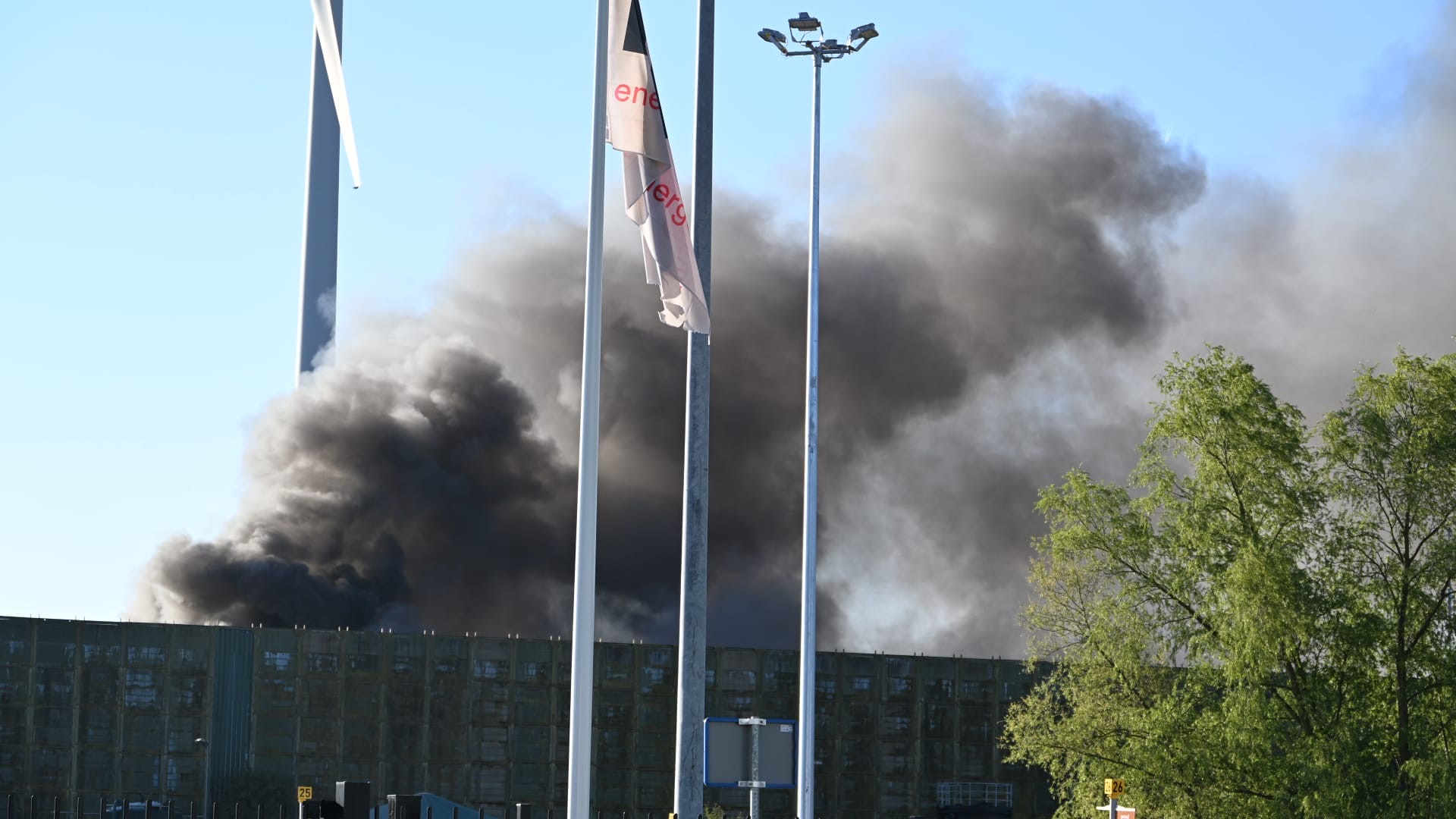 Dikke zwarte rookwolken stijgen op bij een gebouw met windmolens en vlaggen op de voorgrond.