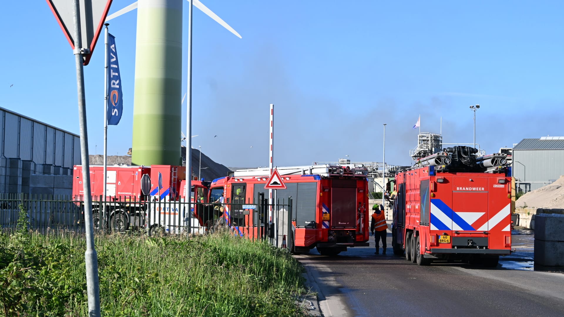 Brandweerwagens bij een industriële locatie met rook op de achtergrond en een groene windmolen aan de zijkant.