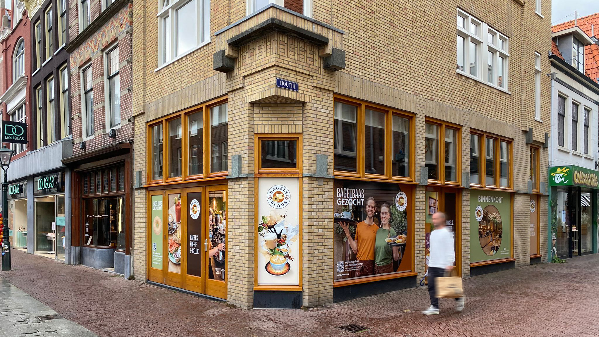 Gevel van een hoekpand met reclame voor Bagels & Beans, met posters die bagels en dranken tonen, en teksten zoals "Bagelbaas gezocht".