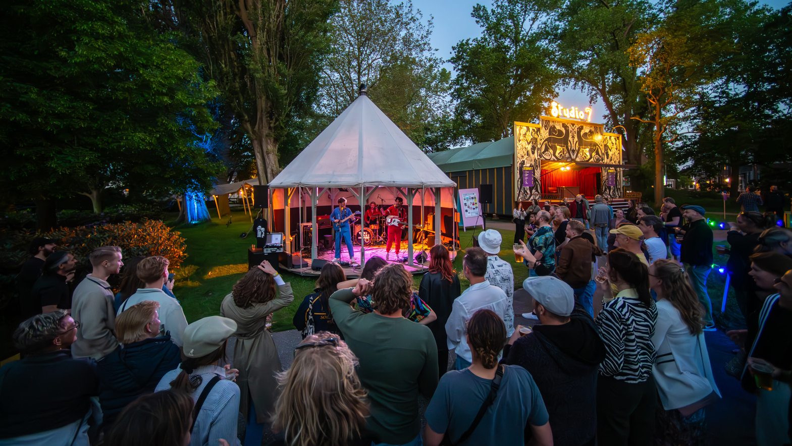 Mensen kijken naar een band die 's avonds optreedt in een witte tent op een festival, met kleurrijke verlichting en bomen op de achtergrond.