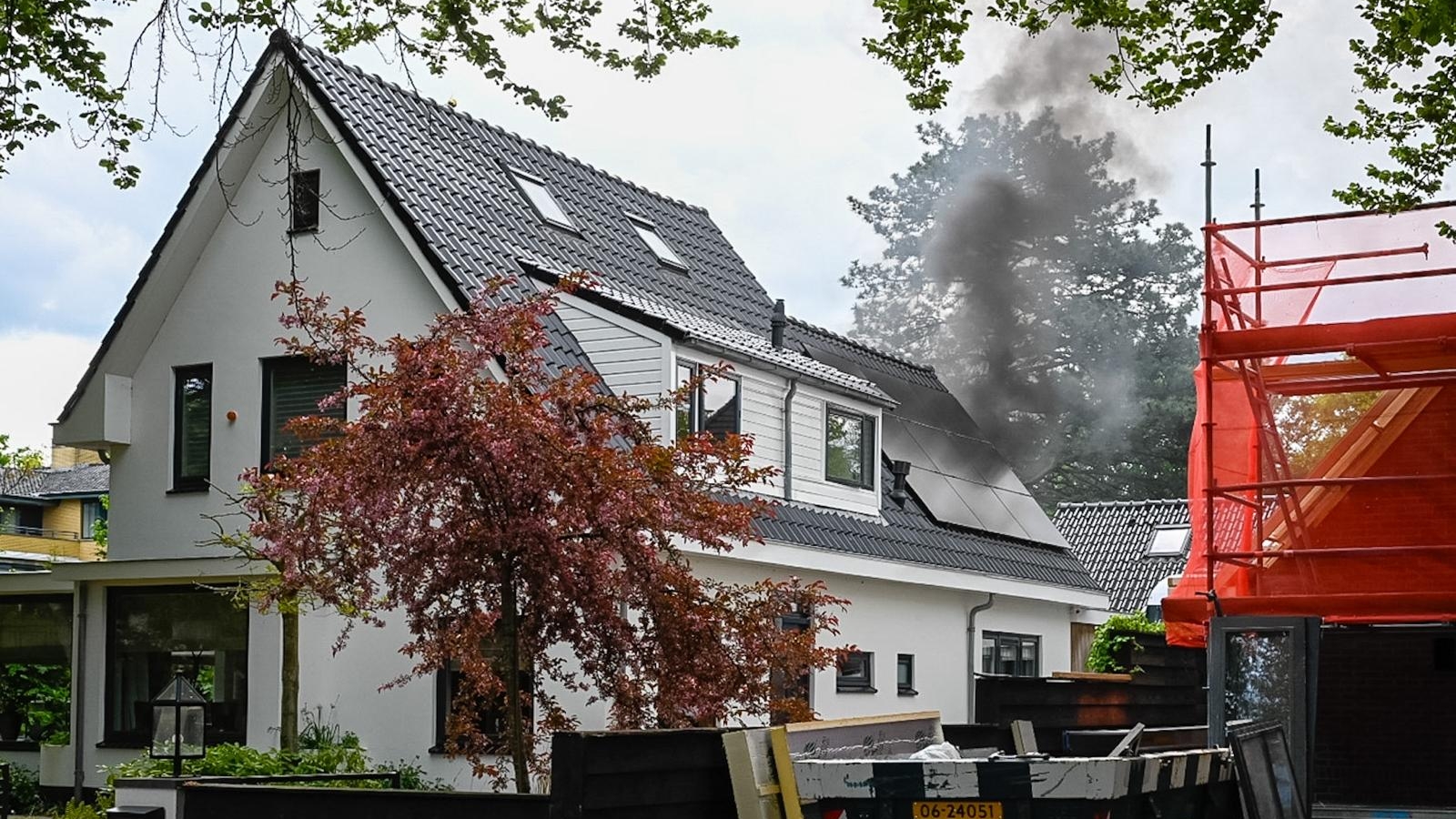 Wit huis met zwarte kozijnen, waar rook uitkomt bij een dakraam; rode bouwsteiger en afvalcontainer op de voorgrond.