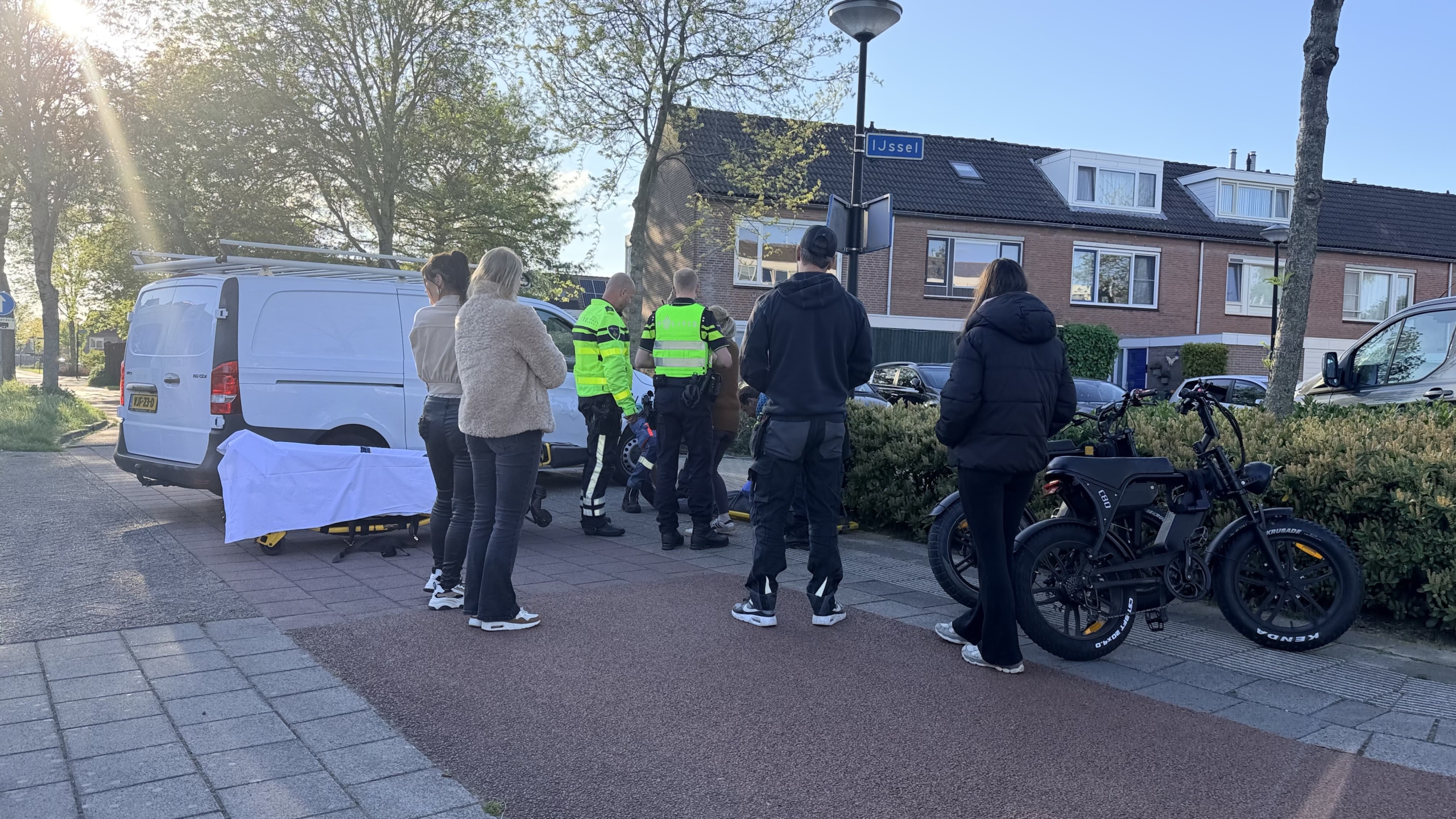 Groep mensen, waaronder politieagenten, verzameld rond een plek op straat met een bestelwagen en enkele elektrische fietsen op de achtergrond.
