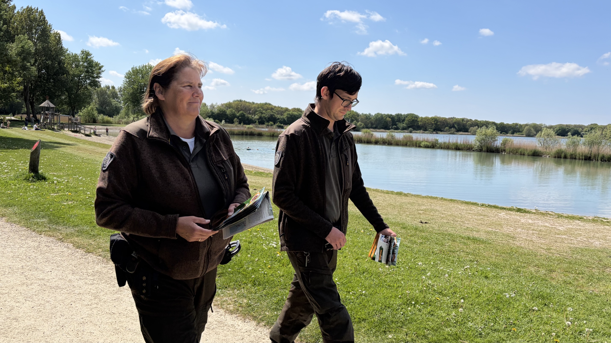 Twee mensen in uniform lopen langs een meer op een zonnige dag, met folders in hun handen.
