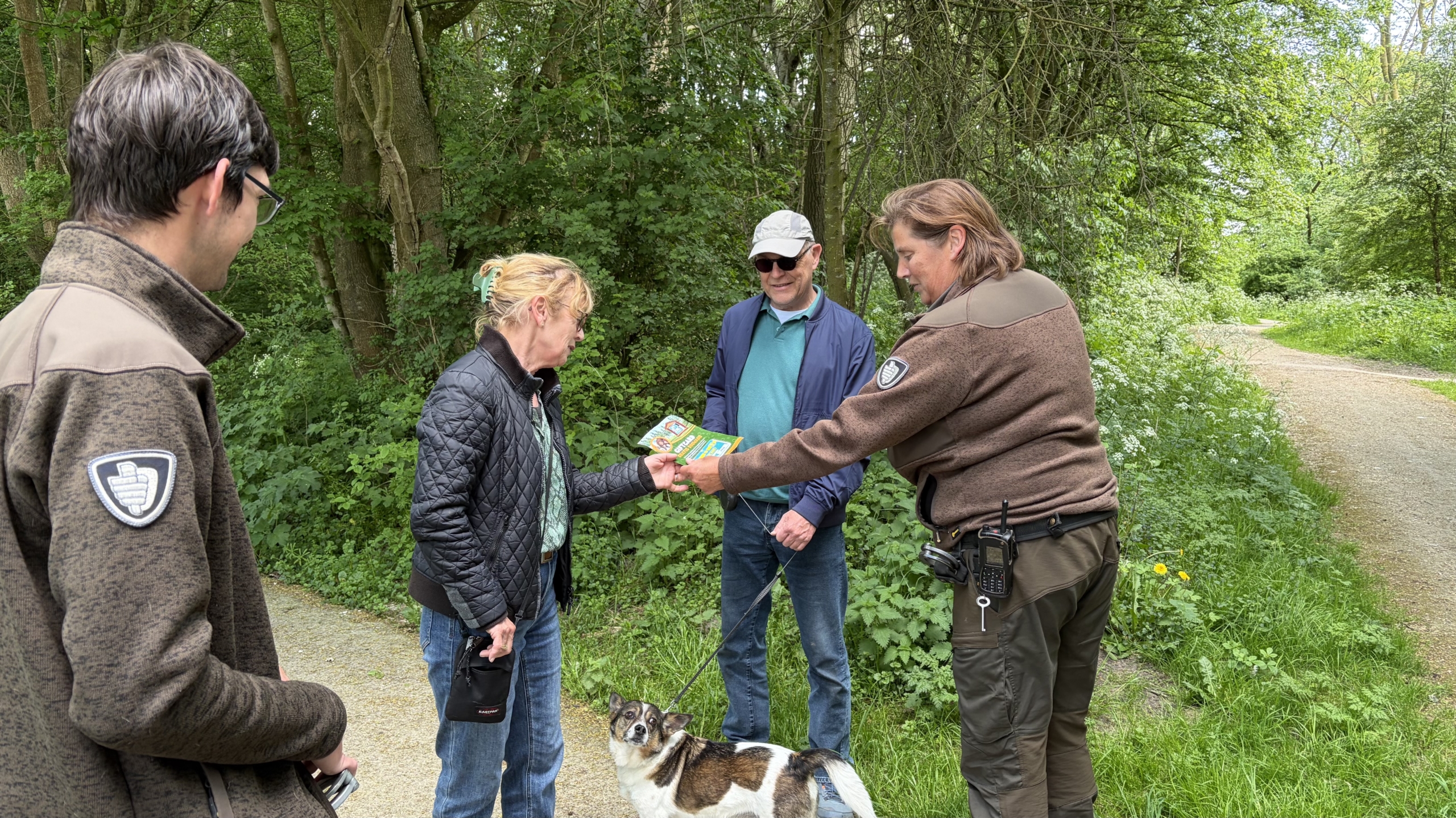 Vier mensen staan in een bos, waarbij een vrouw een folder ontvangt van een man in een groen uniform. Er is een hond aanwezig.