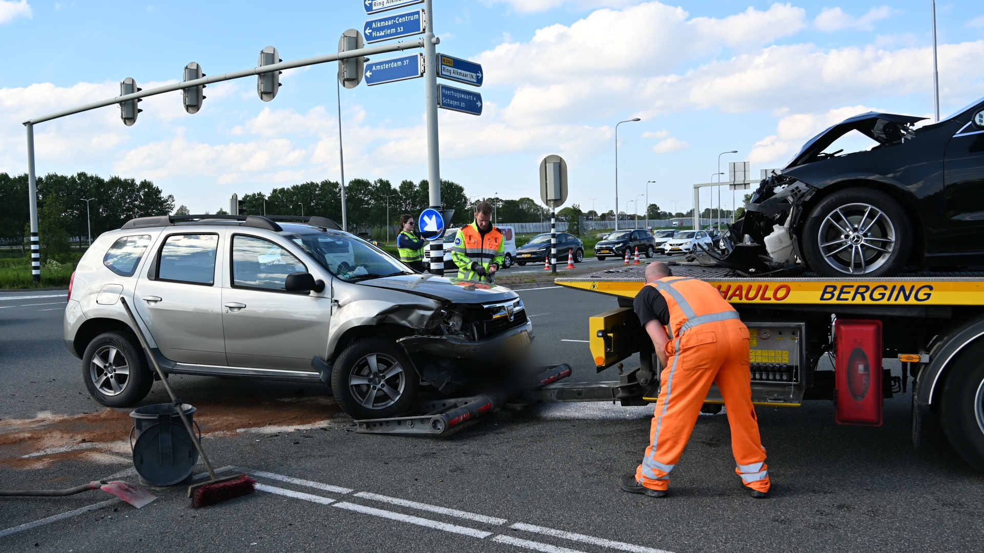Twee auto's met schade na een botsing worden afgevoerd door een bergingsvoertuig; er zijn mensen in veiligheidskleding aanwezig.