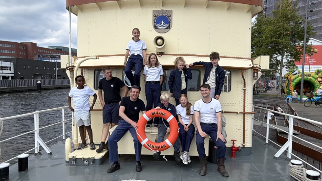 Een groep mensen, voornamelijk in uniform, poseert op een schip met een reddingsboei waarop "Bulgia Alkmaar" staat.
