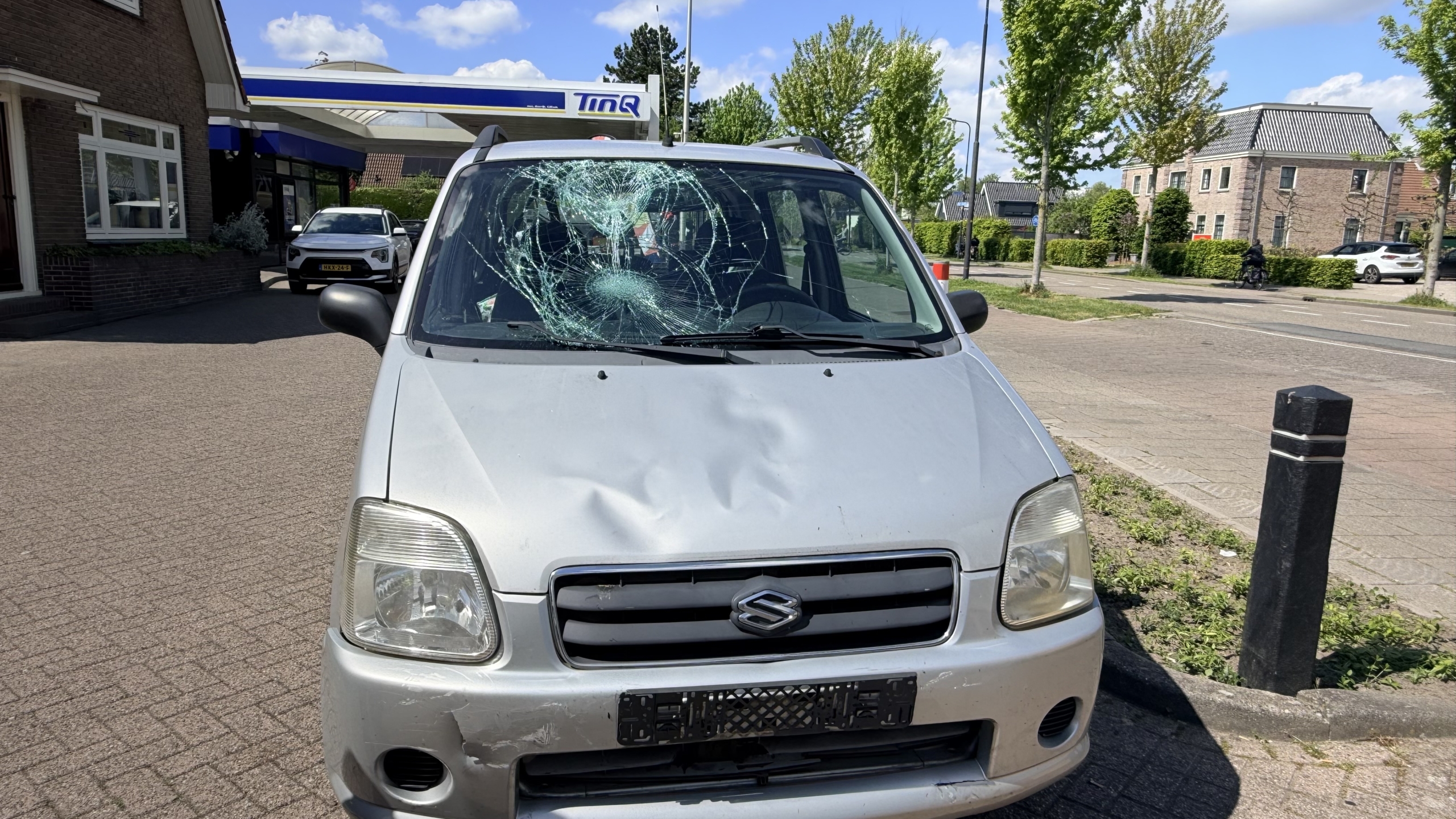 Auto met ingedeukt voorpaneel en gebarsten voorruit, geparkeerd bij een tankstation op een zonnige dag.