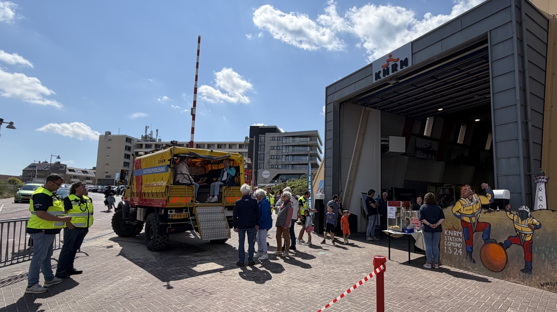 Mensen staan buiten een KNRM-station naast een voertuig met open achterdeur; het gebouw heeft een open garagedeur en een affiche.