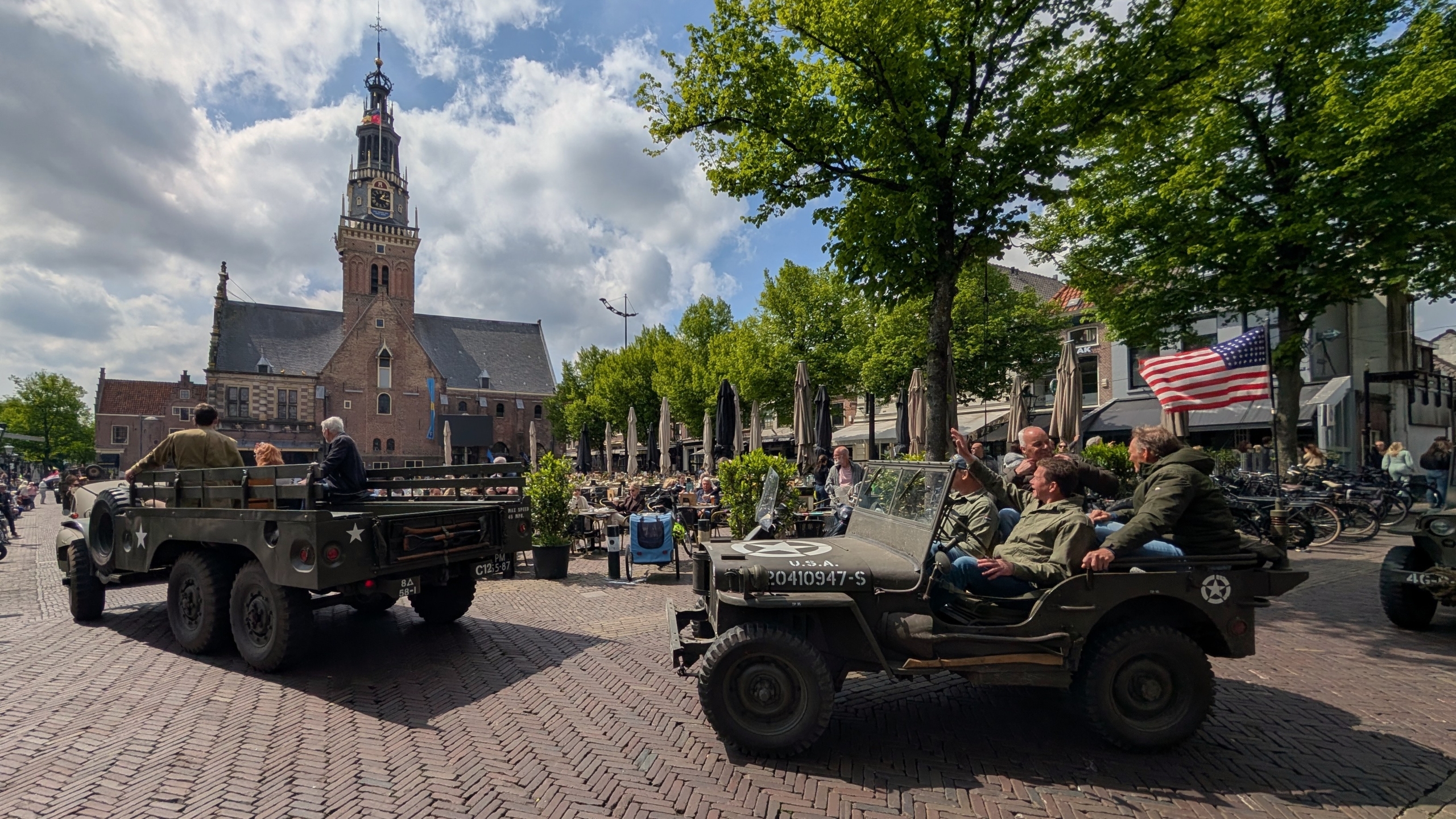 Militaire voertuigen en personen in legerkleding rijden door een plein met een historische kerk op de achtergrond.