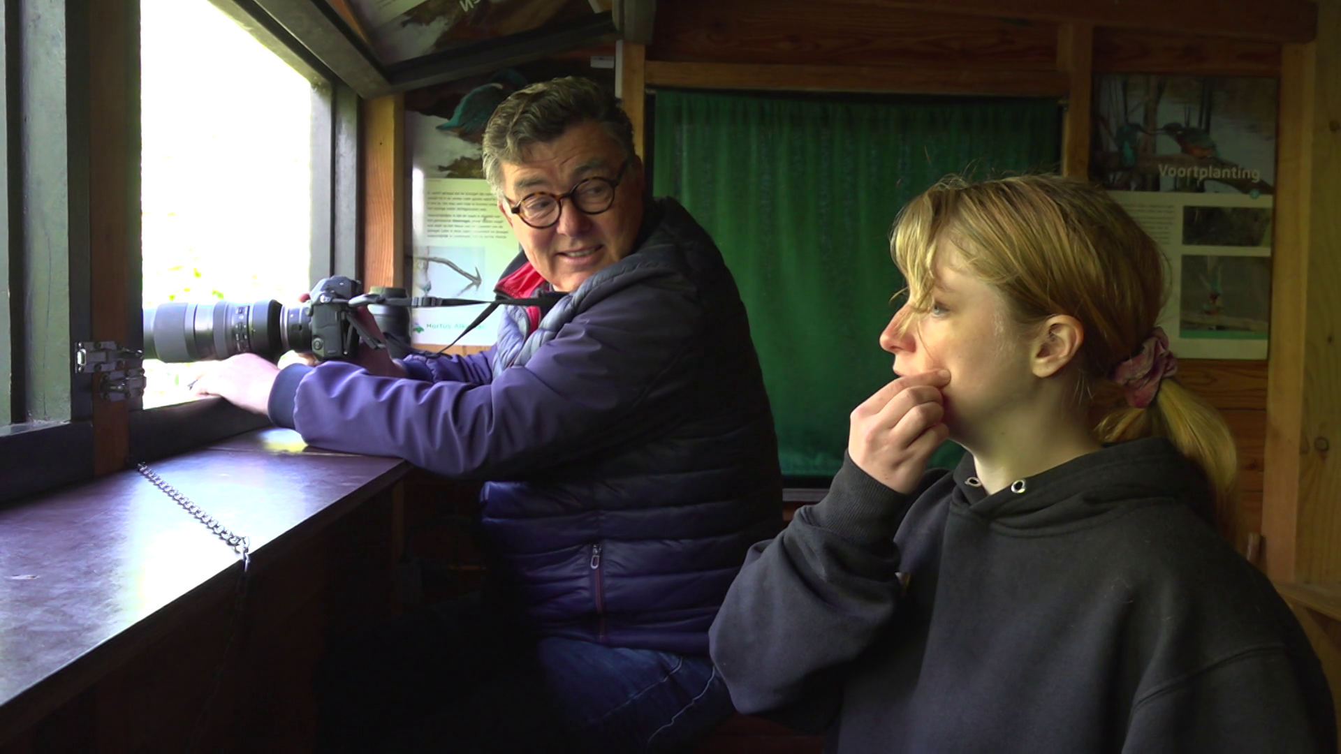 Twee mensen in een vogelkijkhut; één met een camera, de ander kijkt naar buiten.