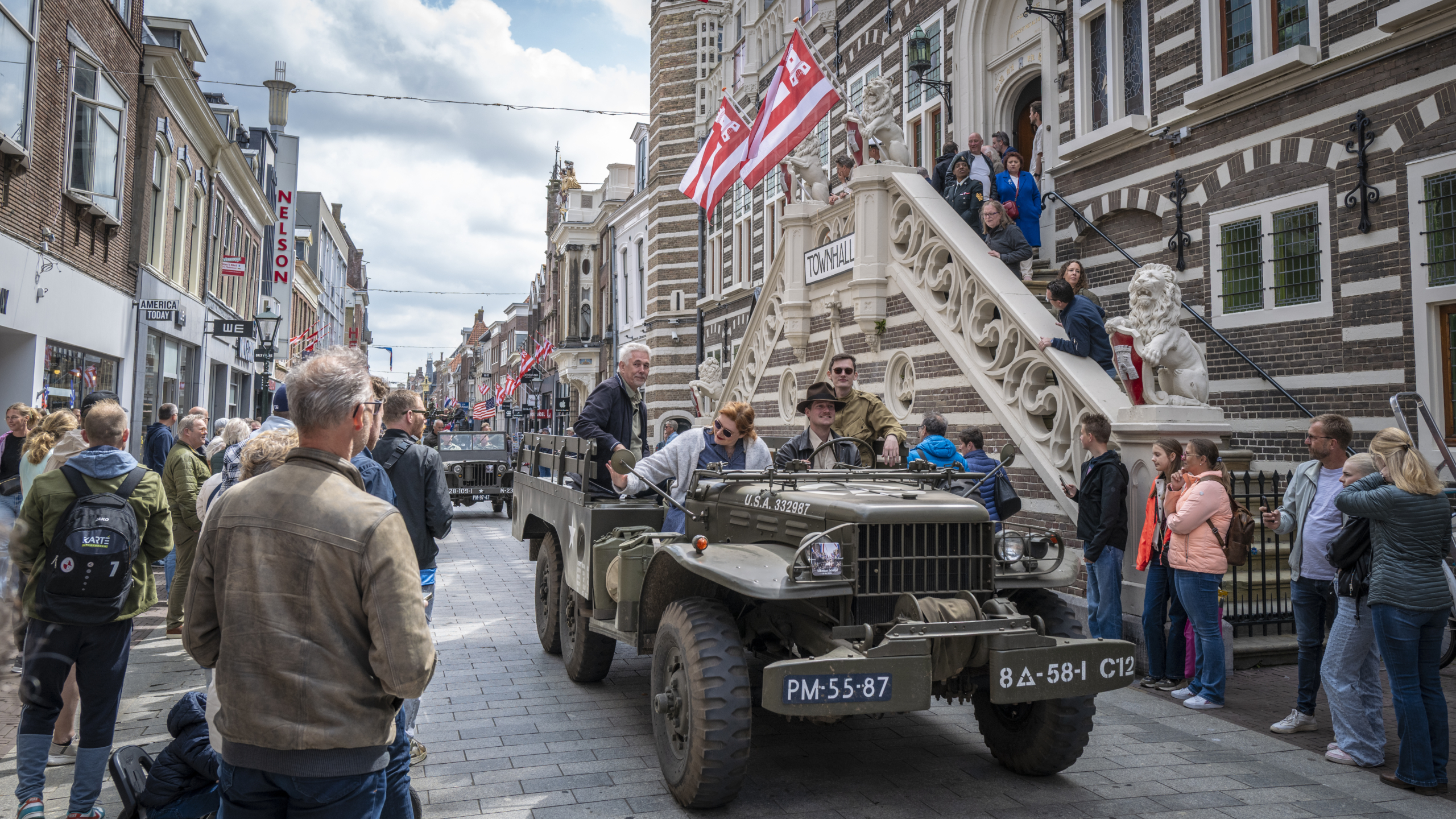 Militair voertuig in parade trekt nieuwsgierige menigte voor een historisch gebouw met Nederlandse vlaggen.