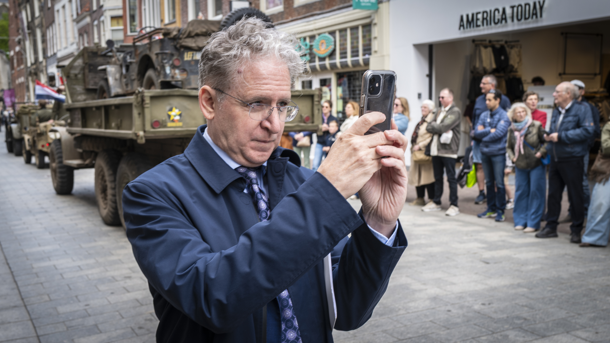 Man maakt een foto met zijn smartphone tijdens een parade met historische legervoertuigen, omringd door kijkers.
