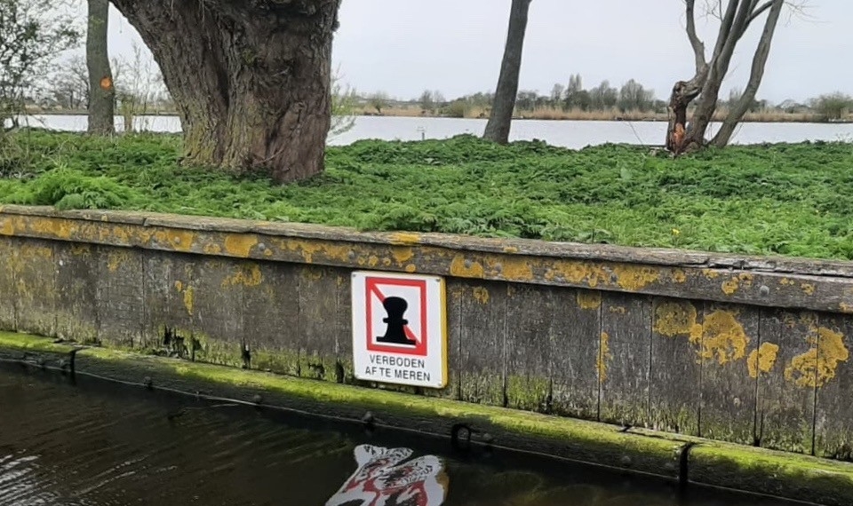 Muur langs een waterkant met een bord "Verboden af te meren" en een achtergrond van groene vegetatie en bomen.
