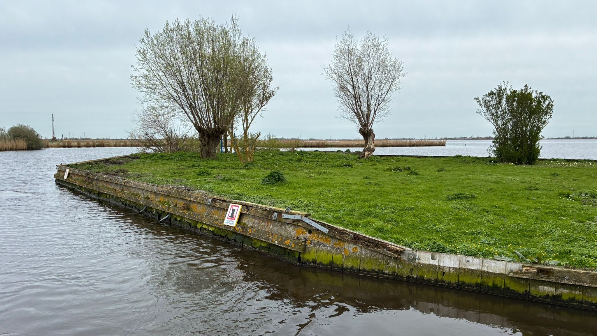 Een klein eilandje met gras en enkele bomen, omgeven door water, met een bordje zichtbaar aan de rand.
