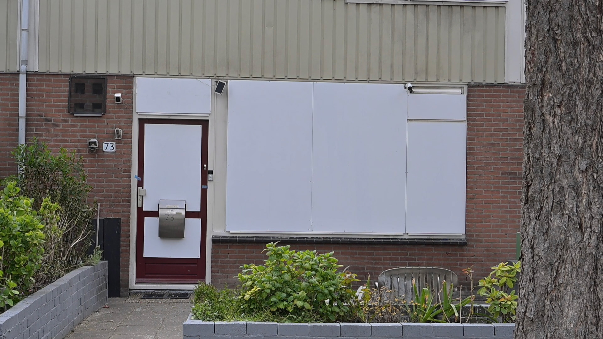 Voorgevel van een huis met een rode deur, dichtgespijkerde ramen, huisnummer 73 en groene planten in de voortuin.