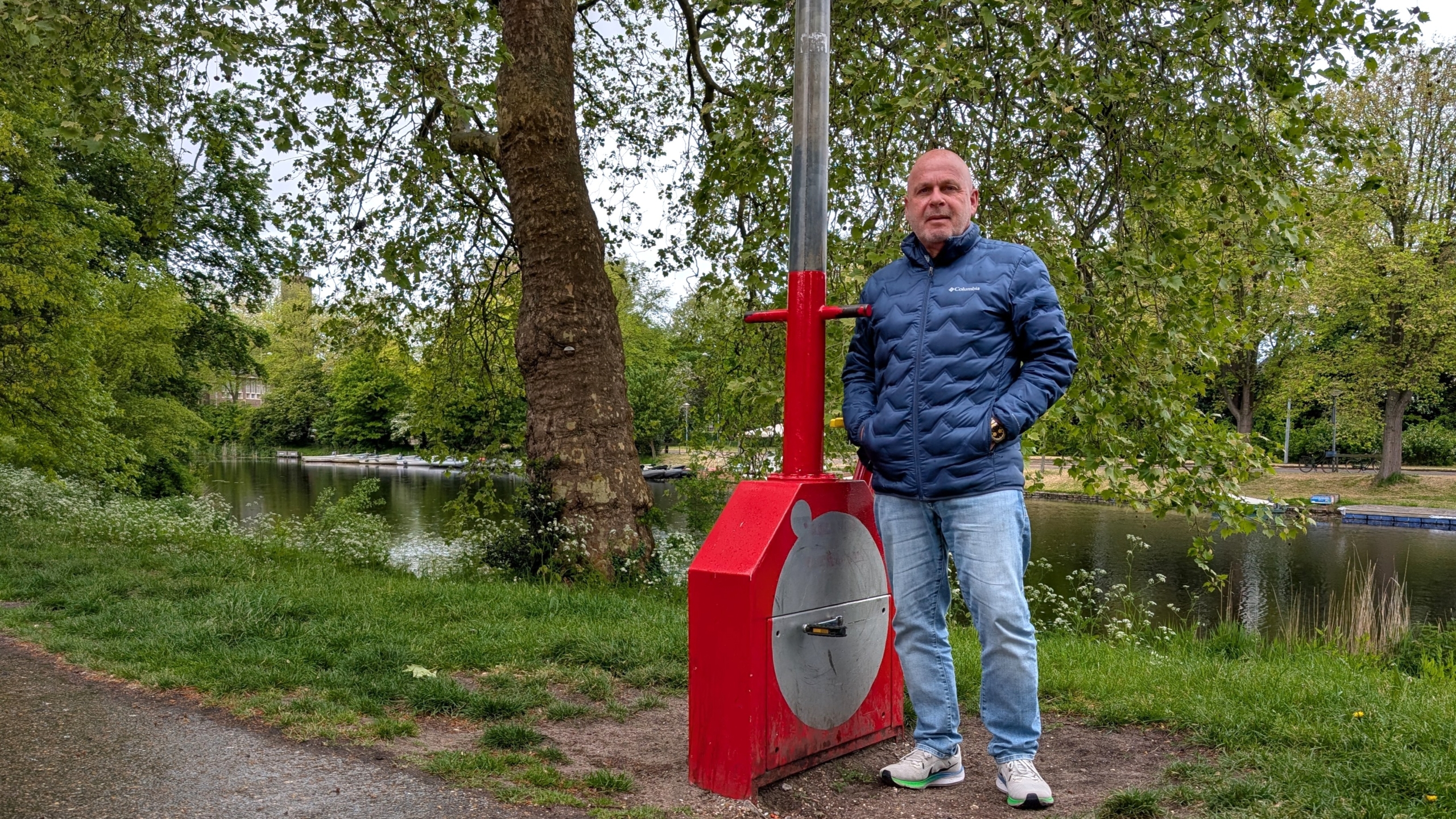Man in blauwe jas staat naast een rood apparaat bij een waterkant met bomen op de achtergrond.