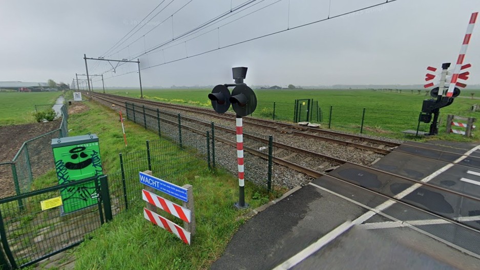 Overweg met een gesloten slagboom, een groen stroomkastje met graffiti en een bord met "WACHT" erop, omgeven door weilanden.