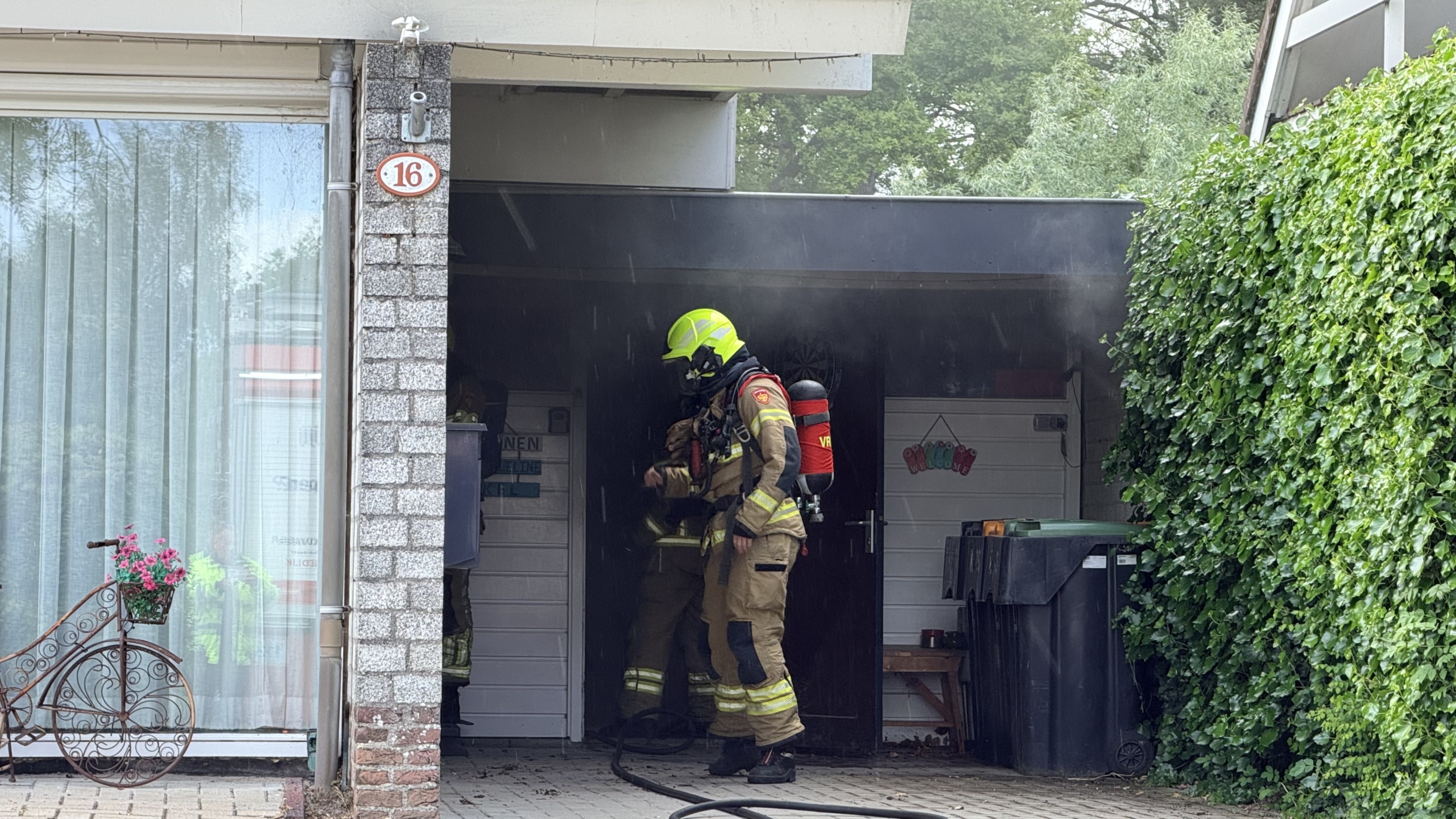 Brandweerman in beschermende kleding betreedt een gebouw met rookontwikkeling naast een woonhuis met een nummer 16.