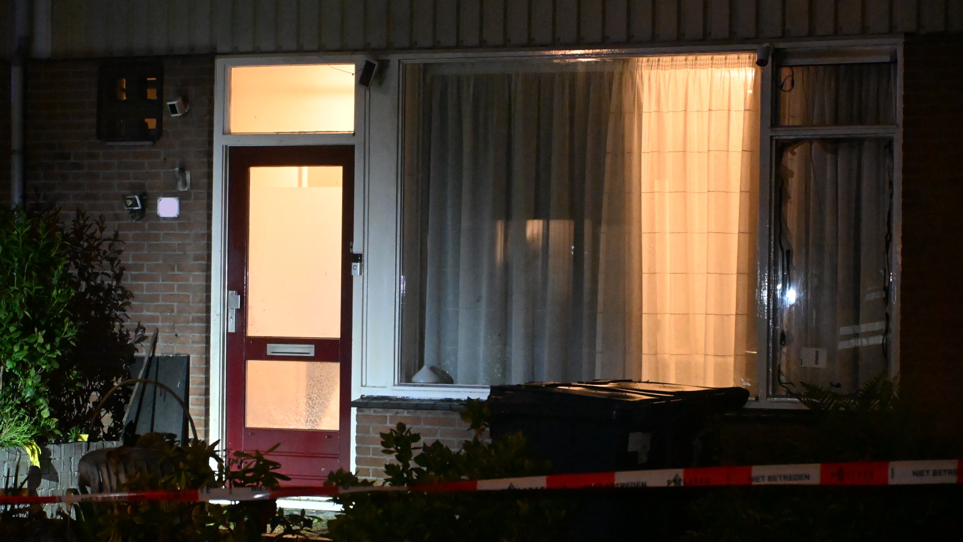 Verlichte voordeur van een huis 's nachts, omgeven door planten en afgesloten met politieafzetting.