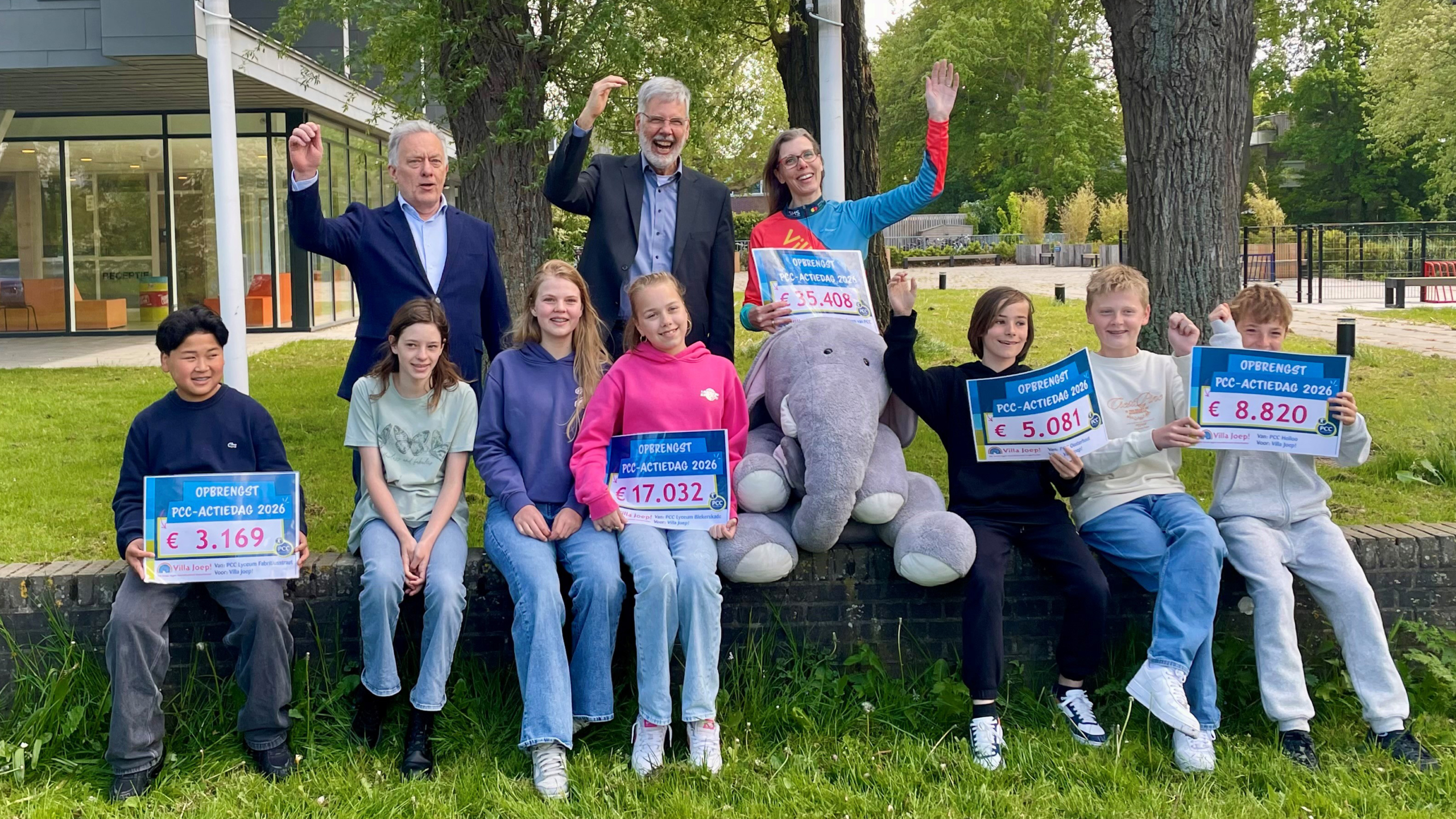 Een groep mensen en kinderen zit buiten op een muurtje, elk met borden die de opbrengst van een inzamelingsactie tonen.