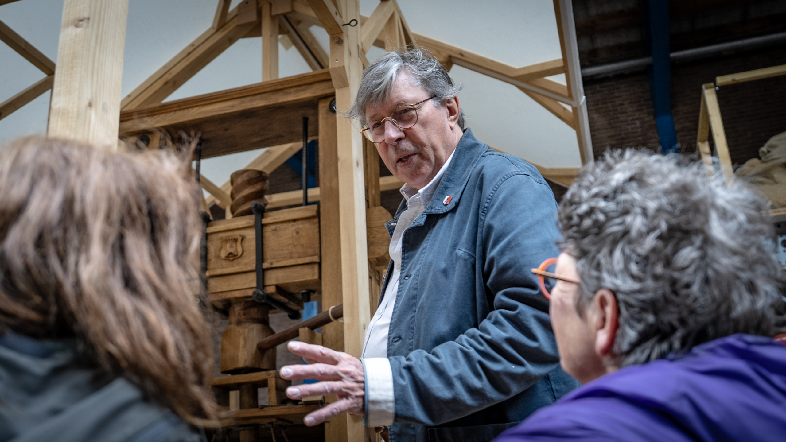 Een man geeft uitleg aan twee mensen in een werkplaats met houten constructies.