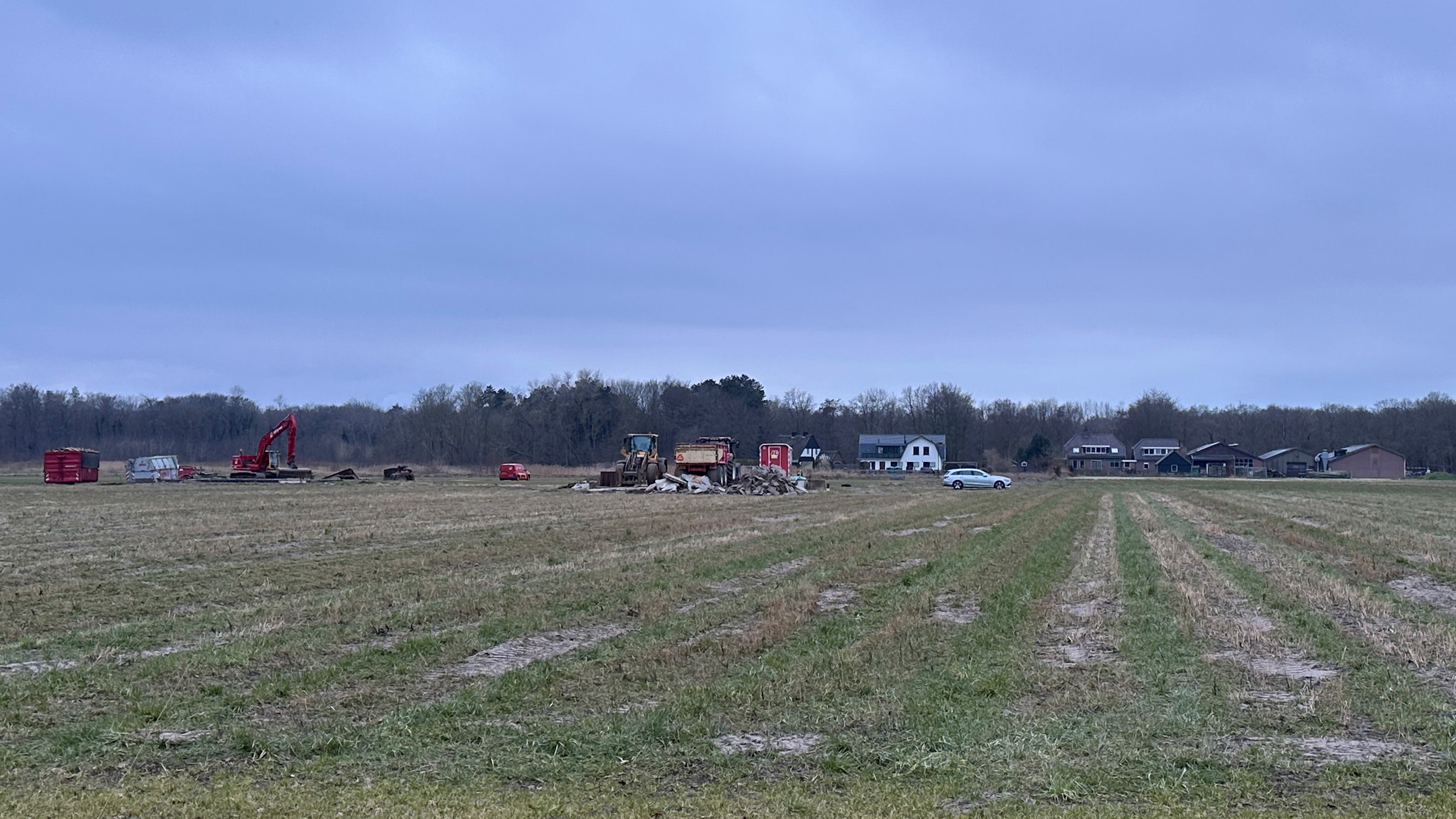 Een veld met bouwmachines, puinhopen en een enkele auto, tegen een achtergrond van woningen en bomen.
