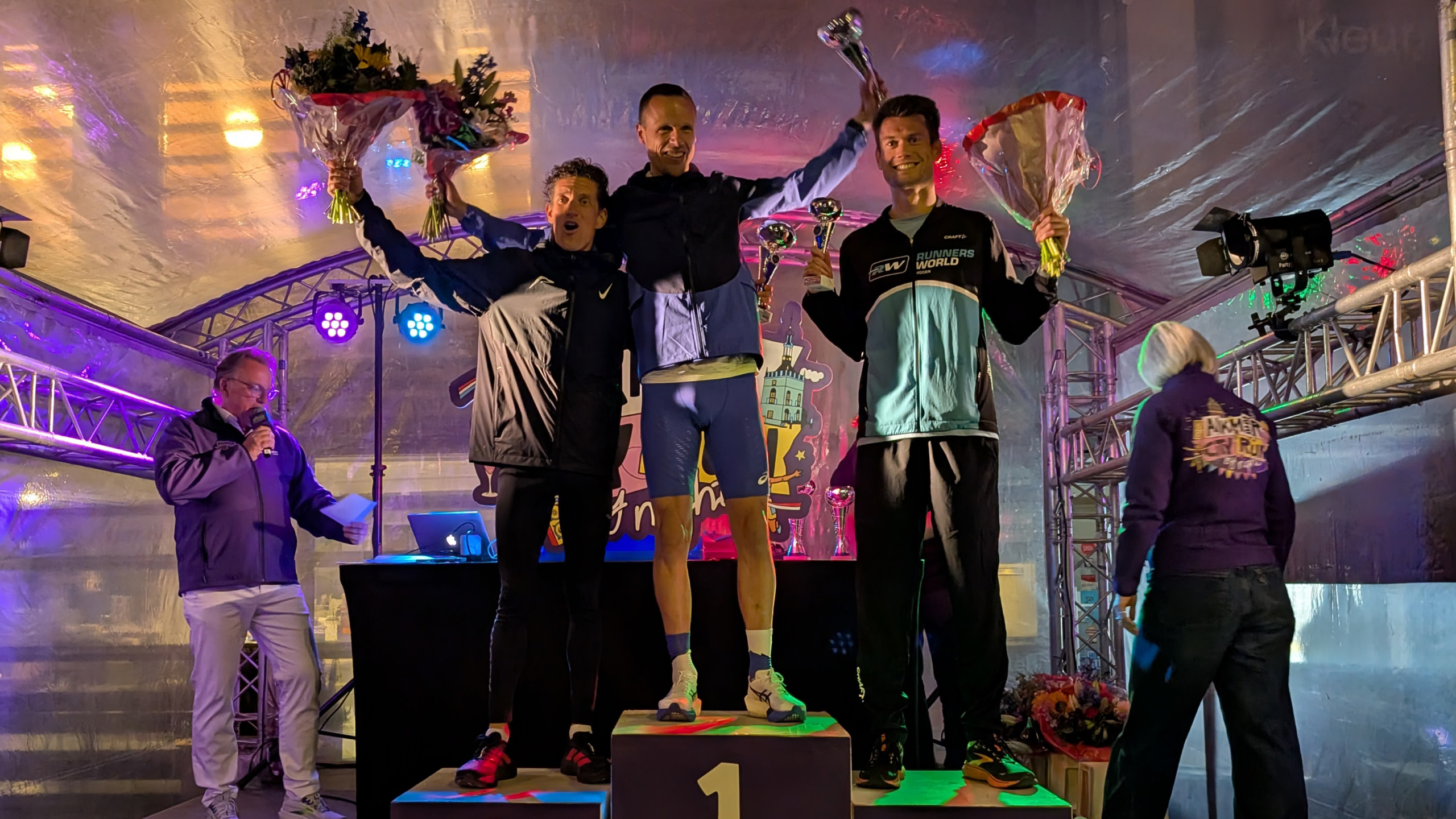 Drie mannen op een podium met bloemen en bekers, onder kleurrijke verlichting op een sportevenement.