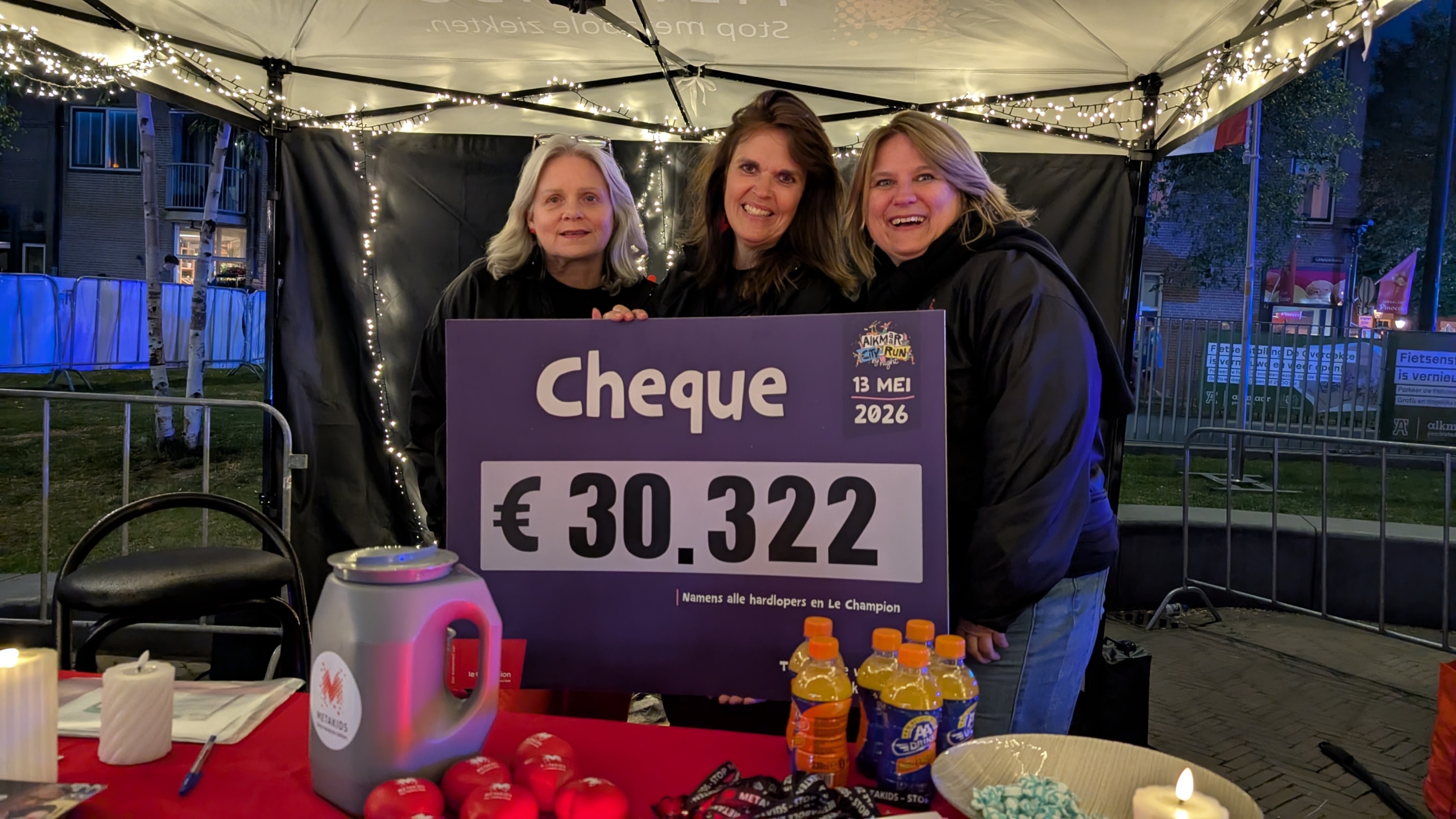 Drie vrouwen poseren onder een tent met een cheque van €30.322, omlijst door kaarsen en drankjes op tafel.