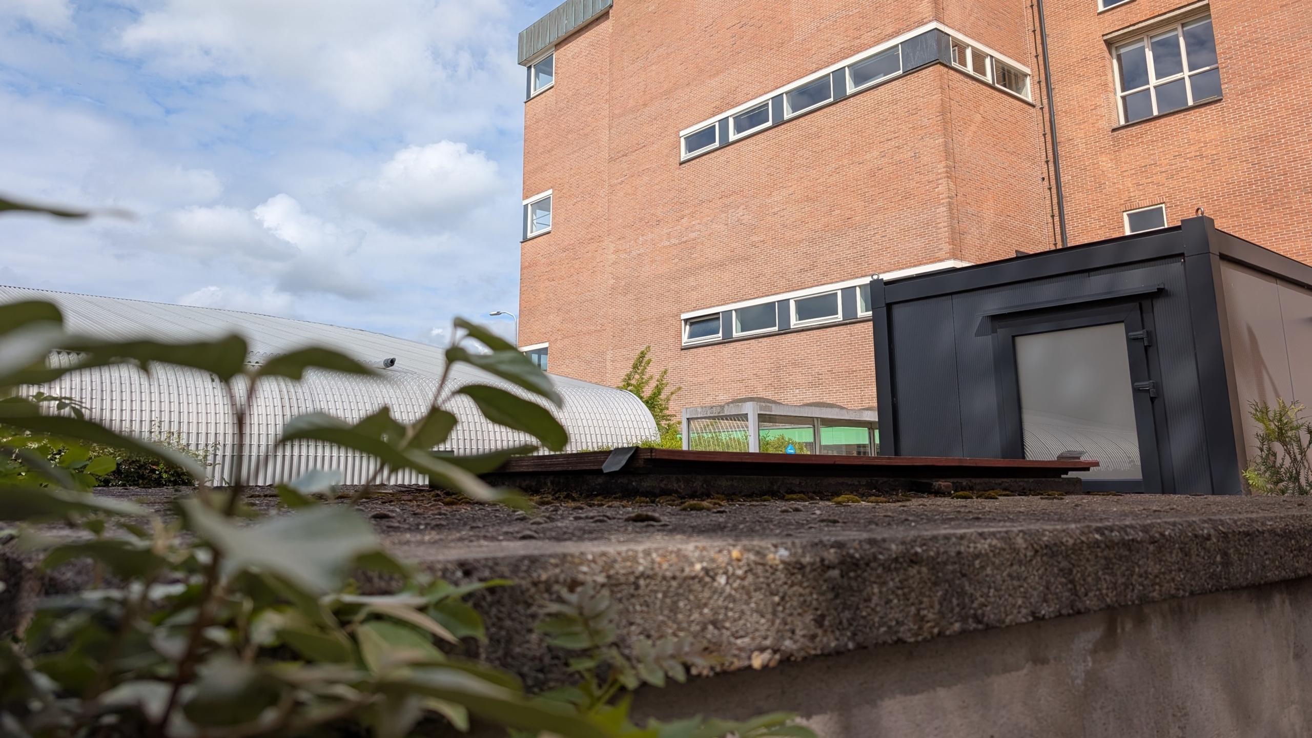 Bakstenen gebouw met ramen, naast een modern zwart gebouw en een gewelfd dak, omgeven door enkele planten op de voorgrond.