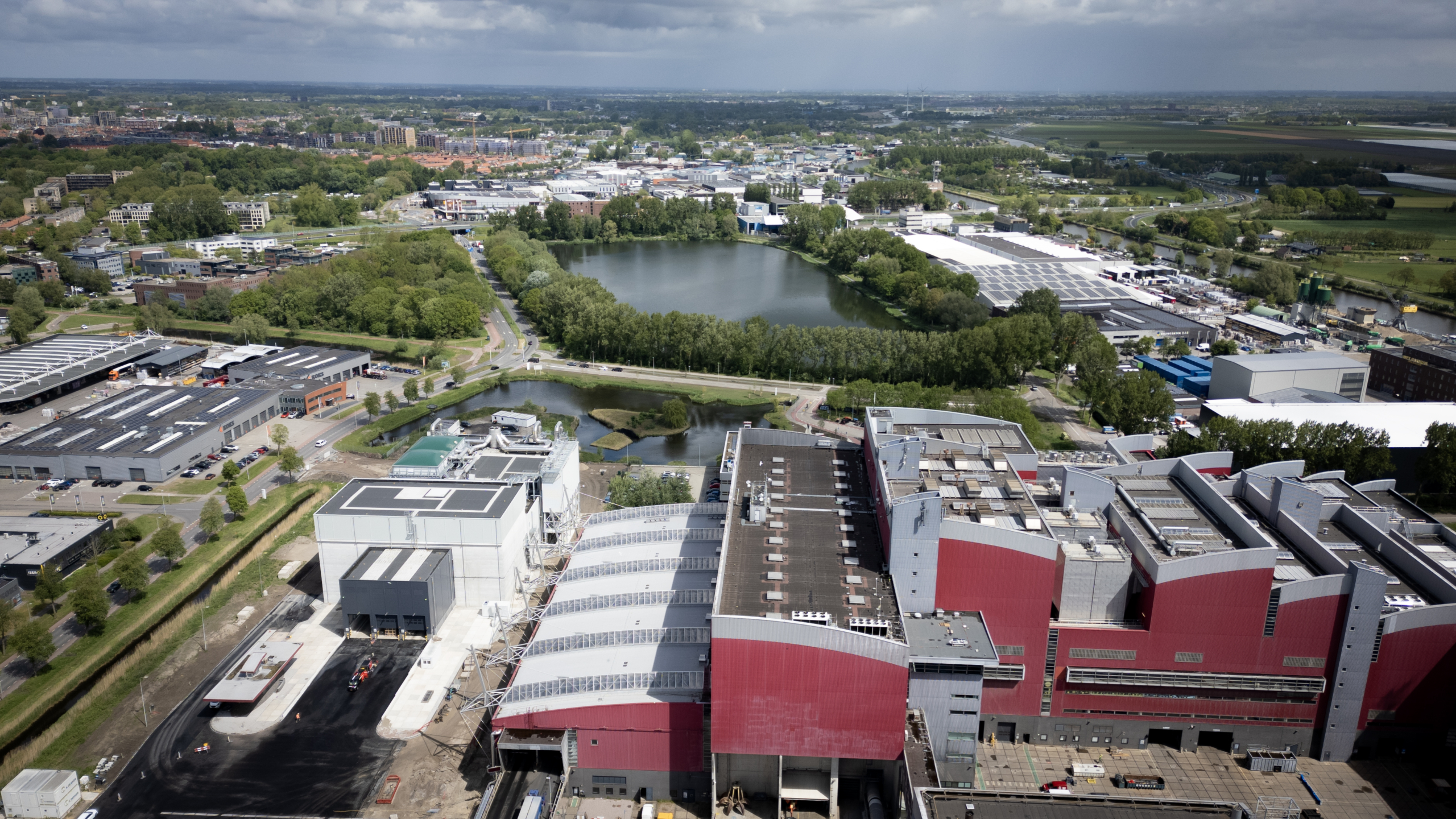 Luchtfoto van een industriegebied met grote rode gebouwen, groene bomen en een meer op de achtergrond.