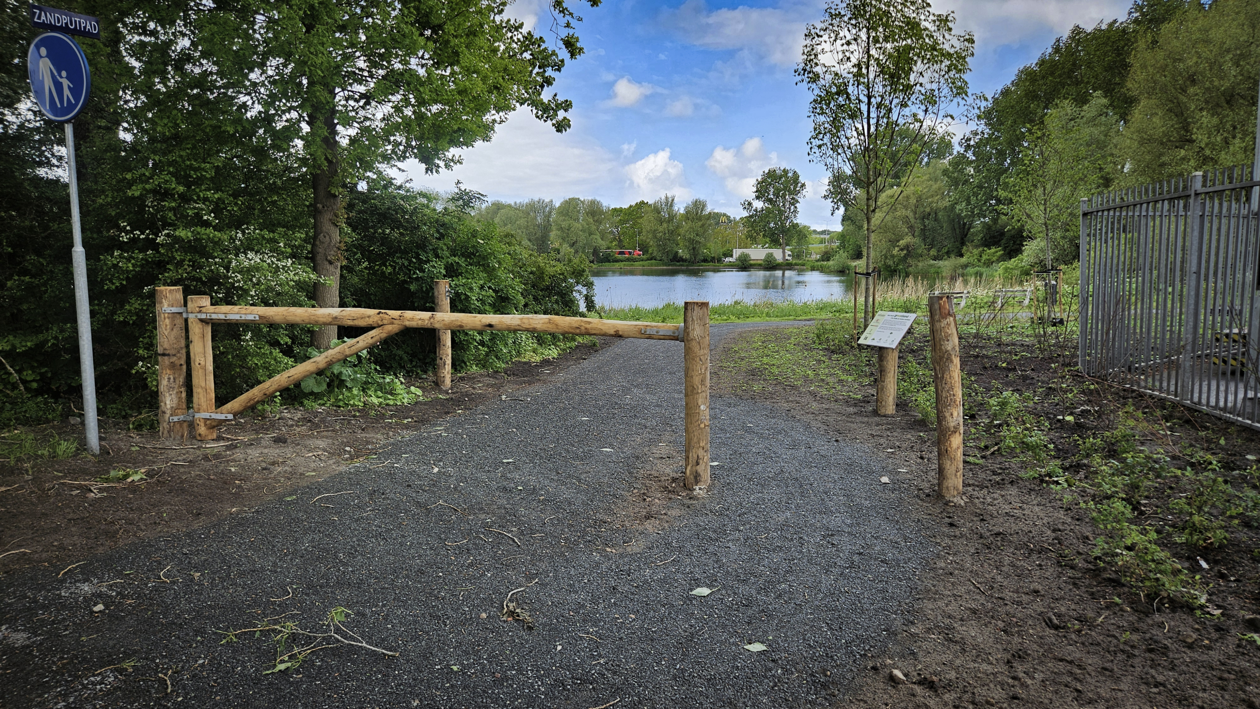 Houten hek met grindpad erachter, omgeven door groen en bomen, naast een bord met tekst "Zandputpad" bij een recreatieplas.