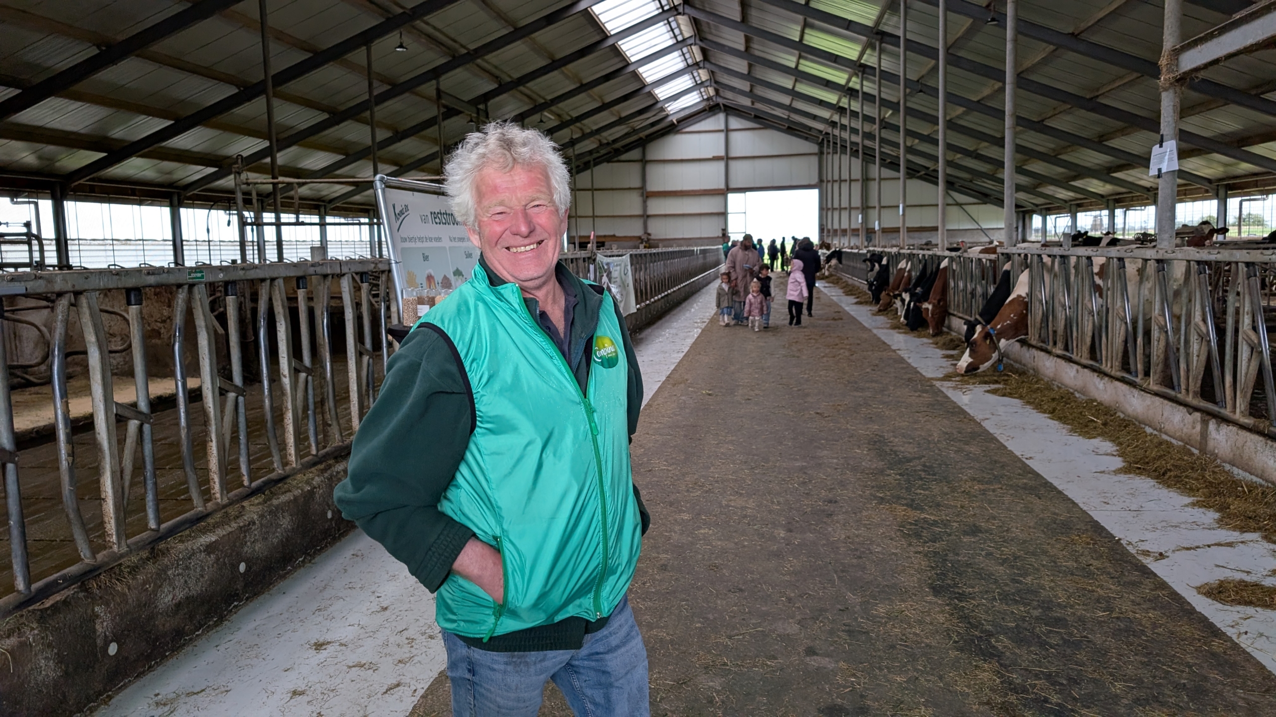 Man in groene jas staat glimlachend in een koeienstal, met kinderen en volwassenen op de achtergrond.