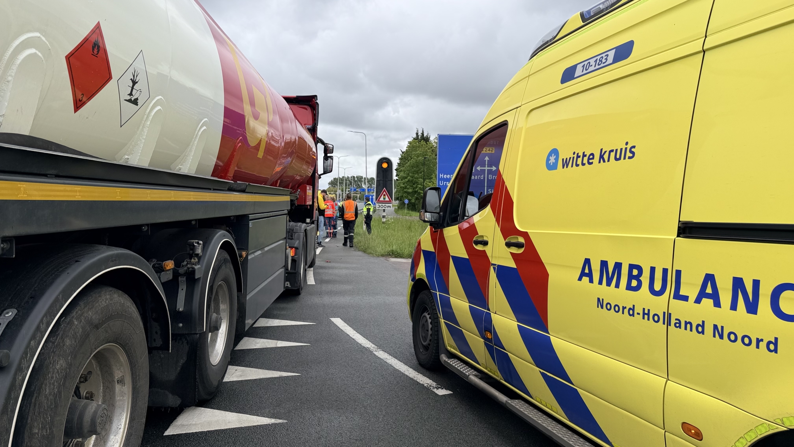 Een gele ambulance en een vrachtwagen zijn gestopt langs de weg, met mensen in veiligheidshesjes verderop.