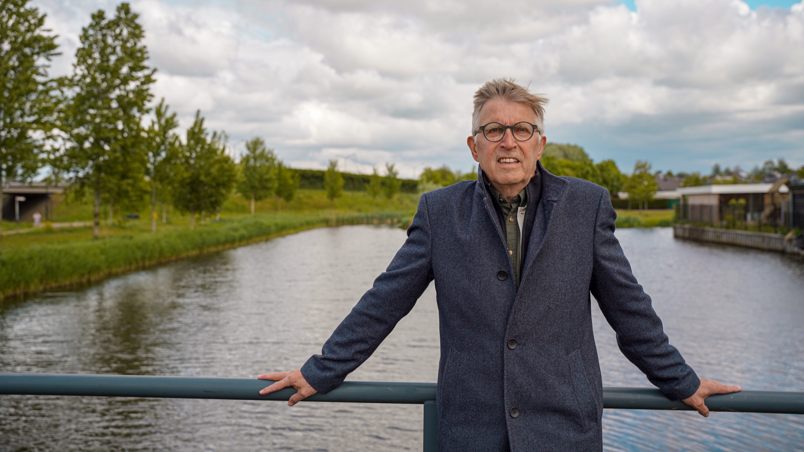 Man met bril en grijze jas leunt tegen een reling, met een kanaal en groene omgeving op de achtergrond.