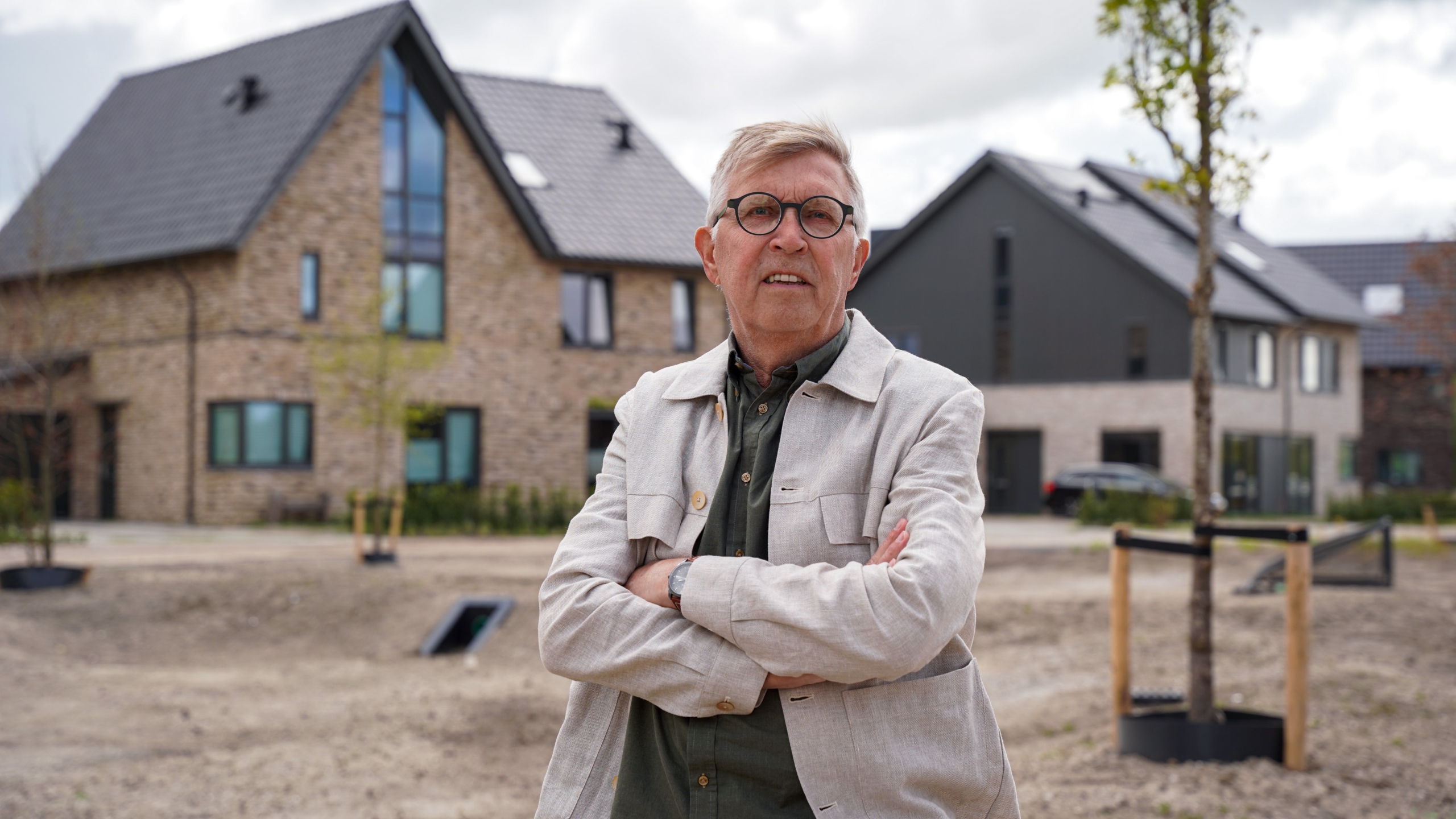 Een man met een bril en grijze jas staat met gekruiste armen voor moderne bakstenen huizen.