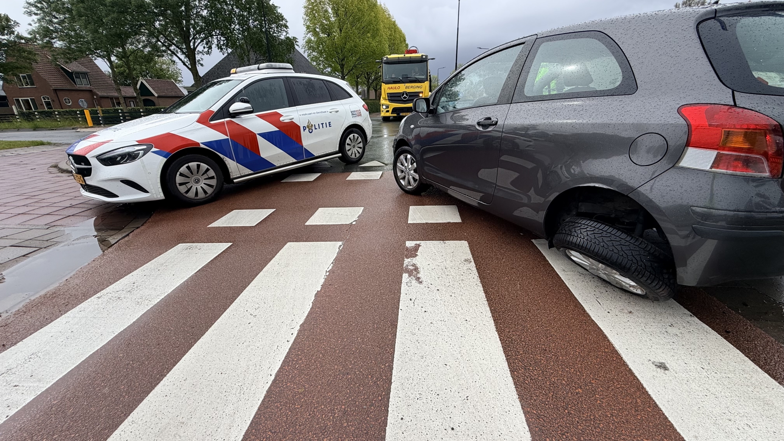 Een politieauto en een grijze auto met een kapot voorwiel staan op een natte weg voor een bergingsvoertuig.