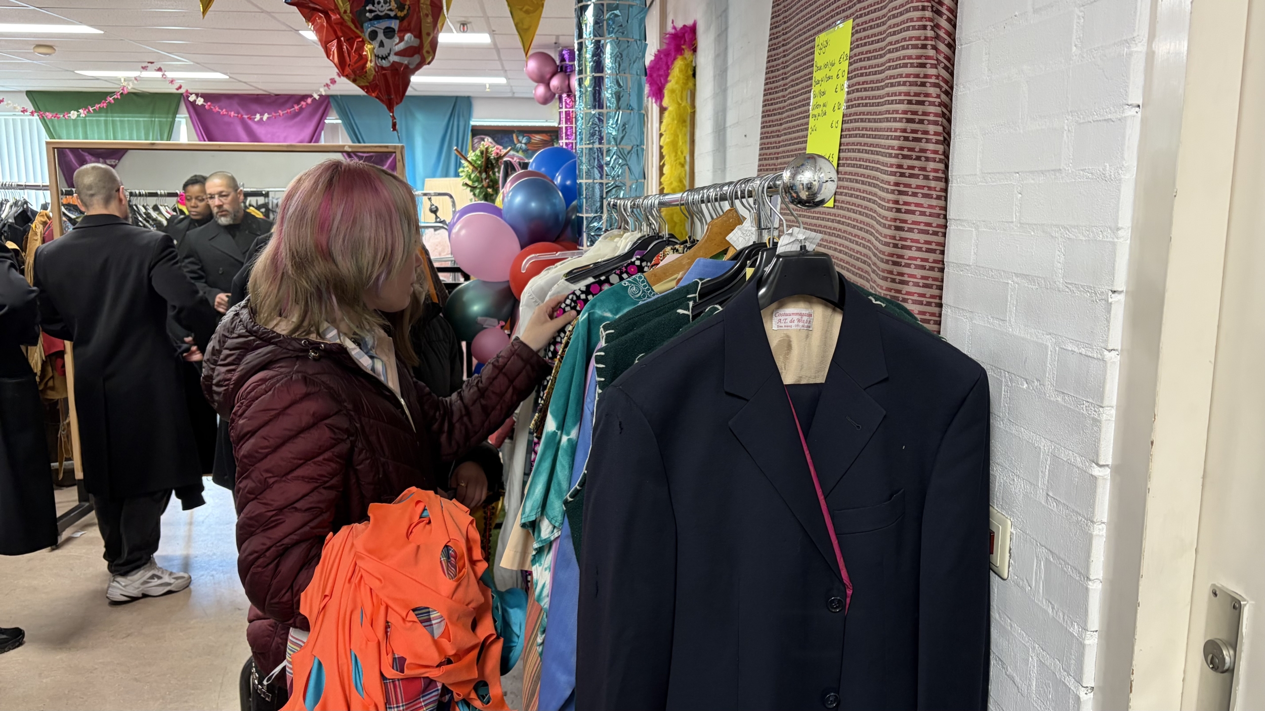 Een vrouw bekijkt kleding aan een rek in een drukke tweedehandswinkel, met feestelijke ballonnen op de achtergrond.