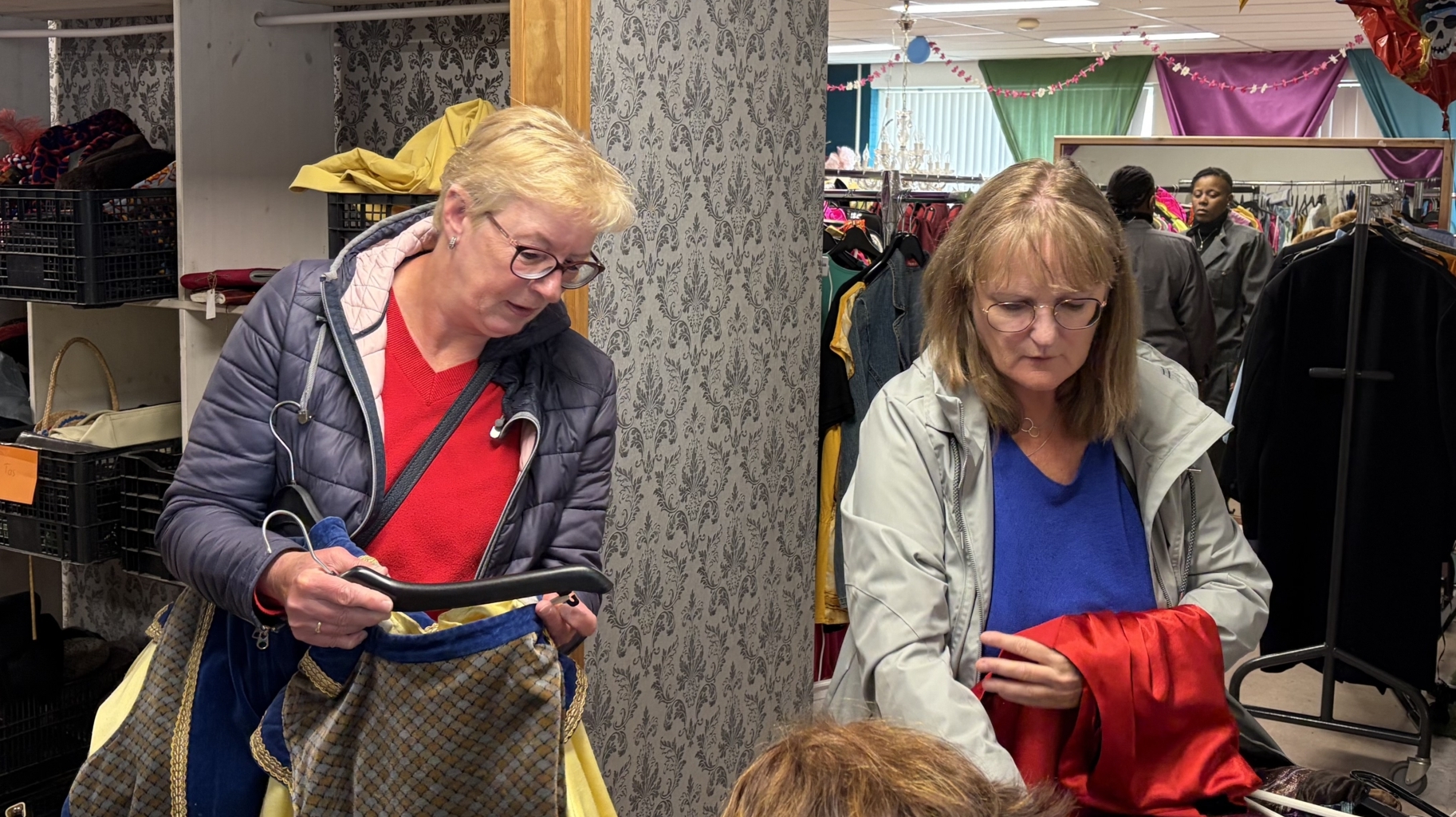 Mensen winkelen in een tweedehands kledingwinkel, omringd door kledingrekken en kleurrijke decoraties.
