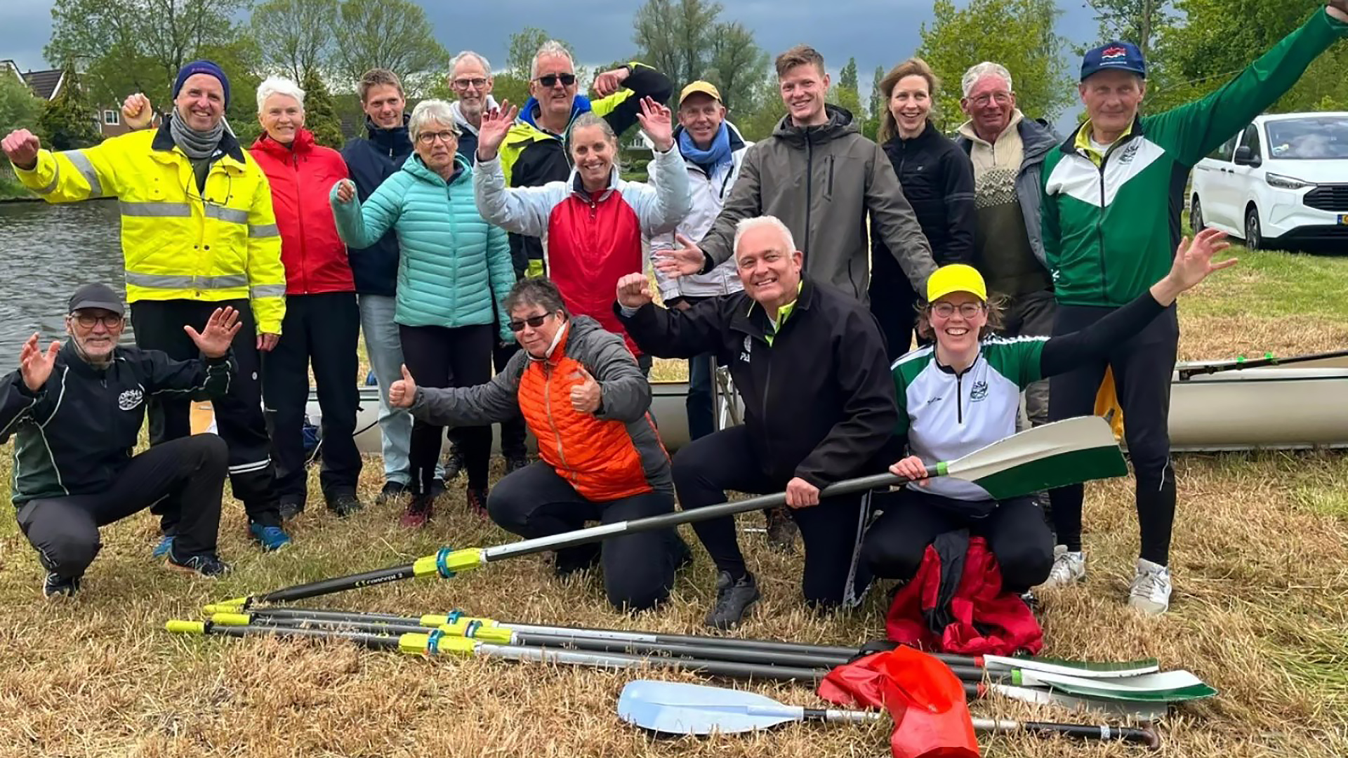 Groep mensen bij een waterkant, lachend en zwaaiend, met roeispanen en een boot op de achtergrond.