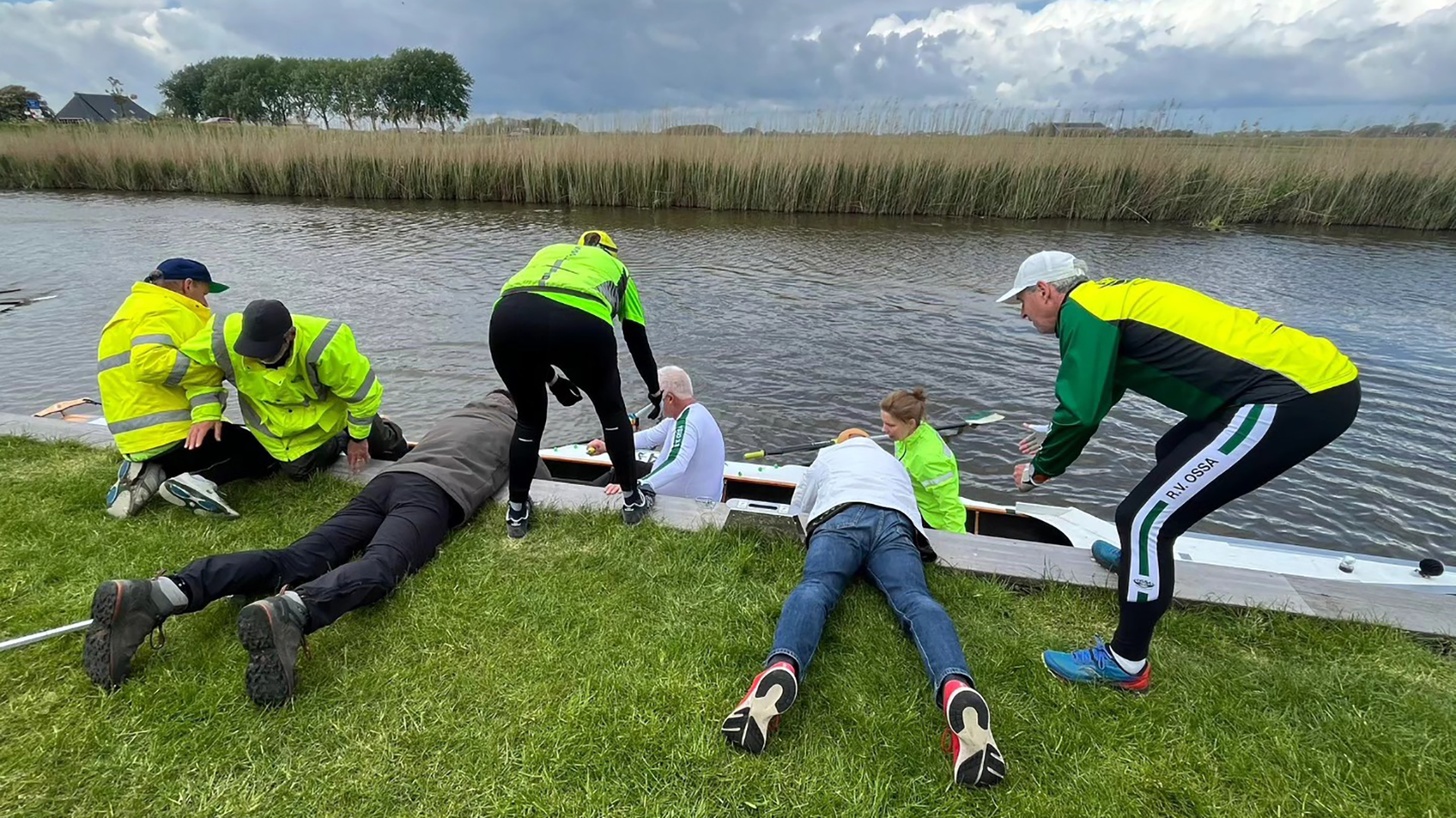 Mensen in felgekleurde kleding helpen een roeiboot langs de waterkant; grassige oever en riet op de achtergrond.