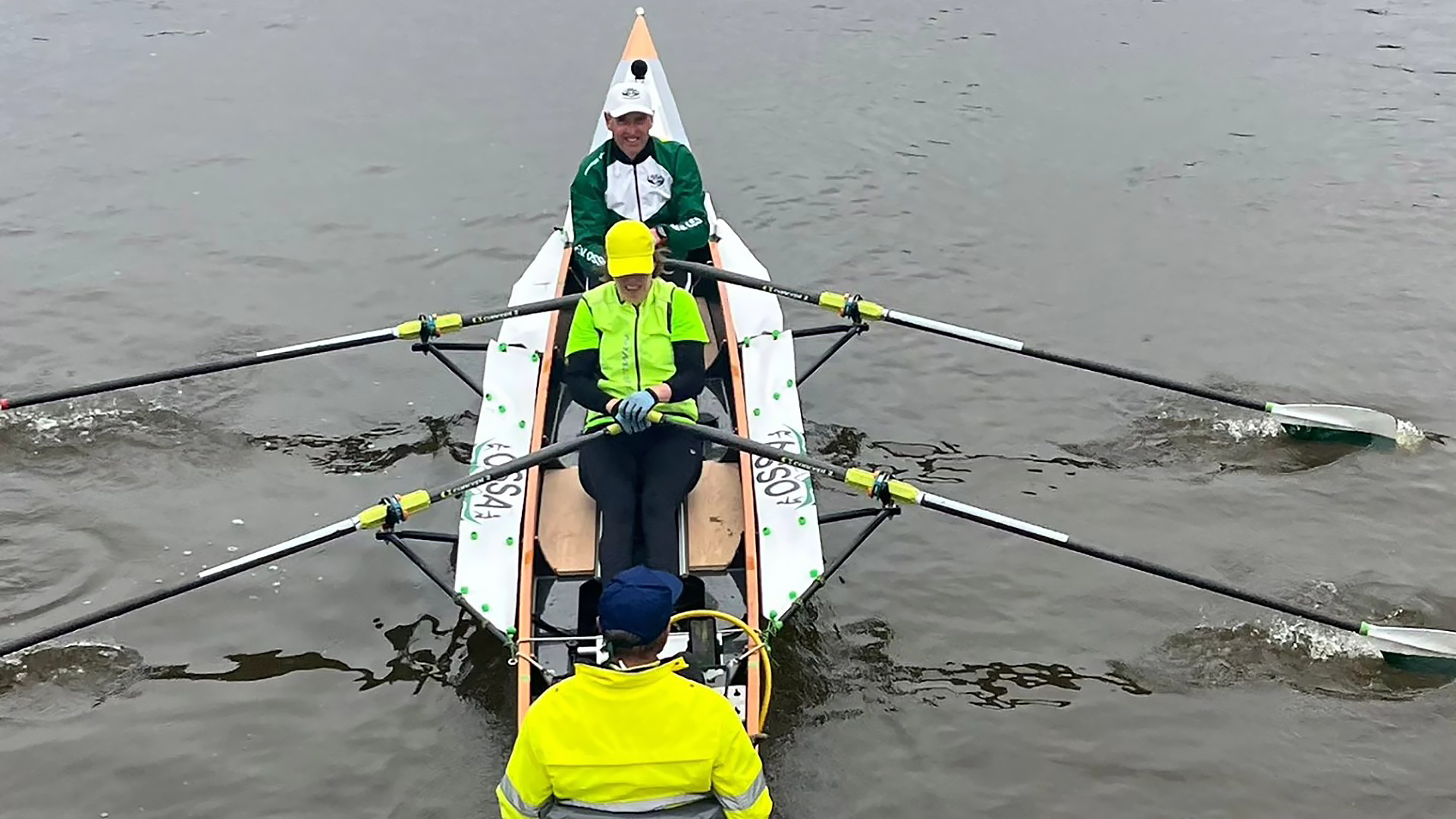 Twee mensen roeien in een dubbele boot met een stuurman op het water, gekleed in felgekleurde kleding.