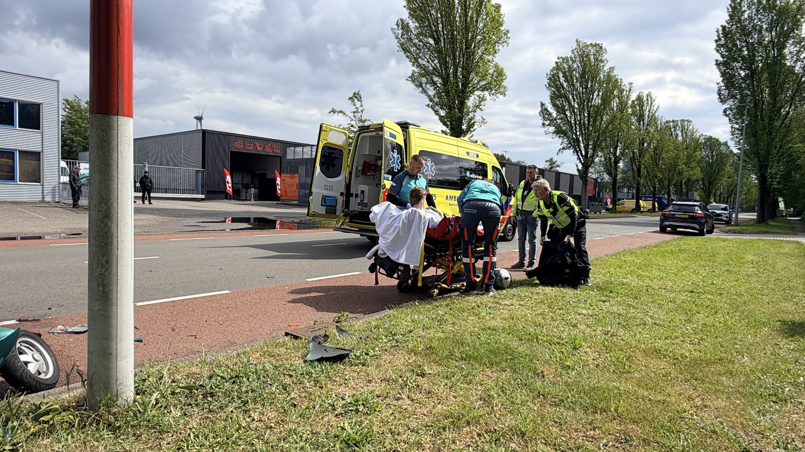 Hulpdiensten helpen een gewonde persoon op een brancard bij een ongeval op straat; een ambulance staat klaar.
