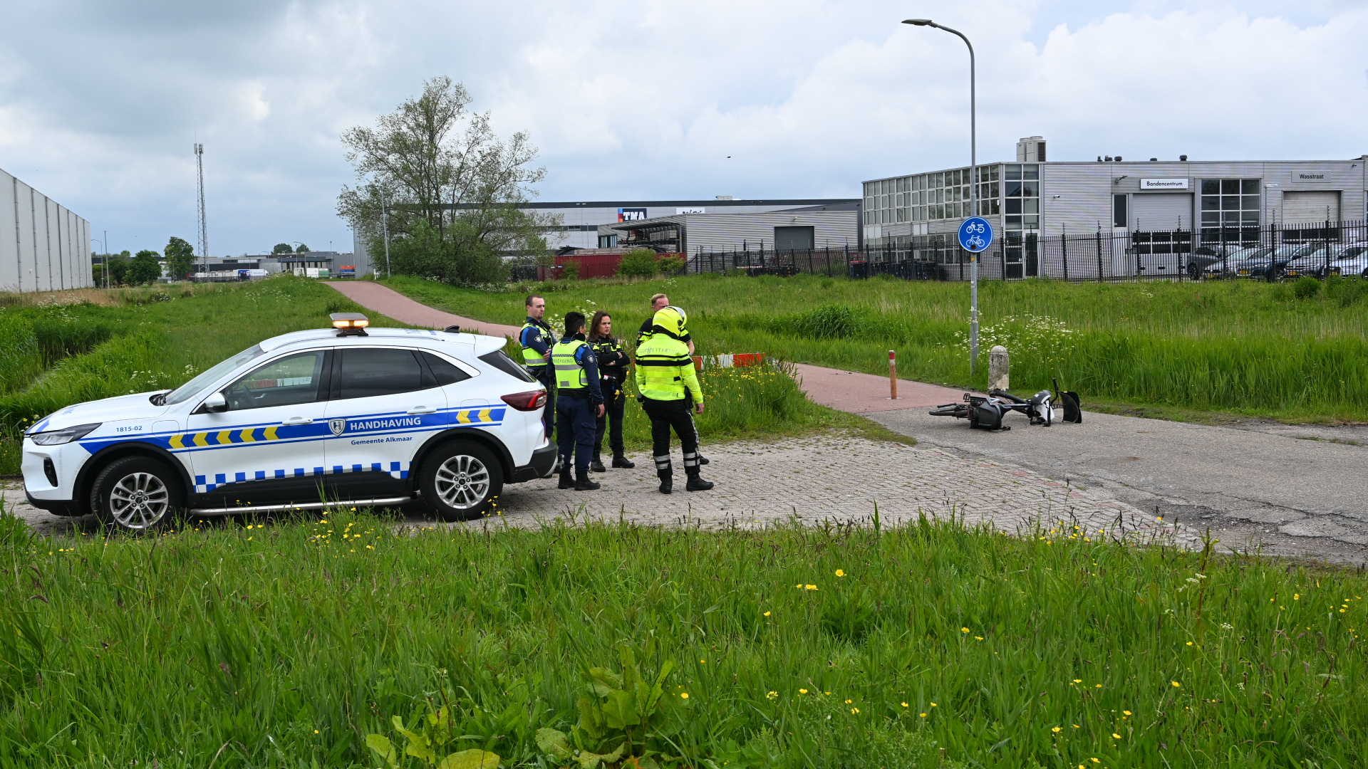 Handhavingsvoertuig bij een fietspad met een omgevallen scooter; handhavers en politie staan naast de auto.