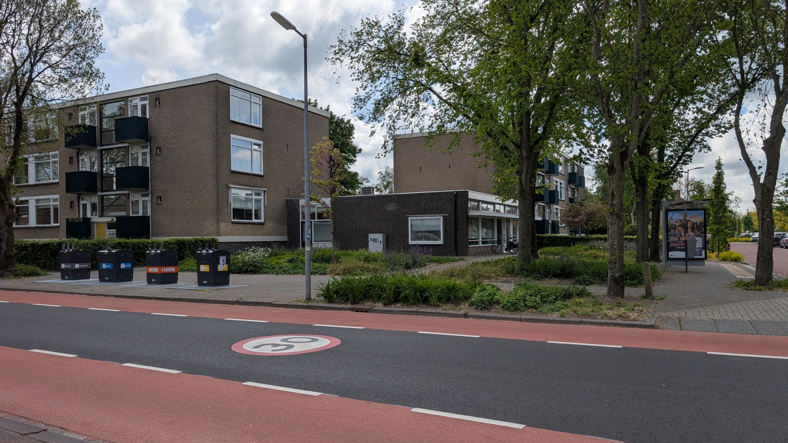 Appartementencomplex naast een weg met bushokje, bomen, en afvalcontainers, onder een bewolkte hemel.