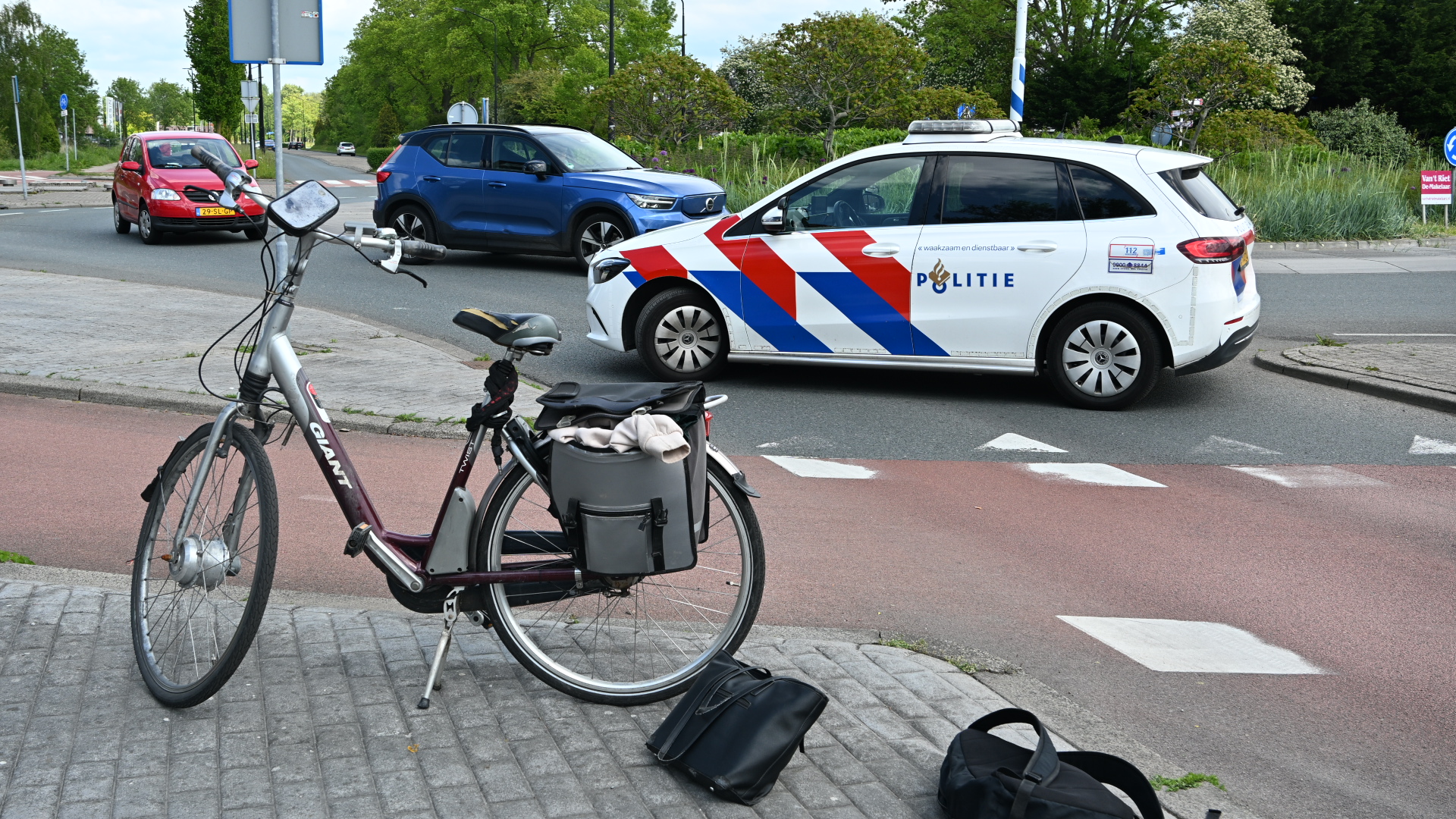 Fiets met tassen geparkeerd bij een rotonde, terwijl een politieauto voorbijrijdt naast twee andere auto's.