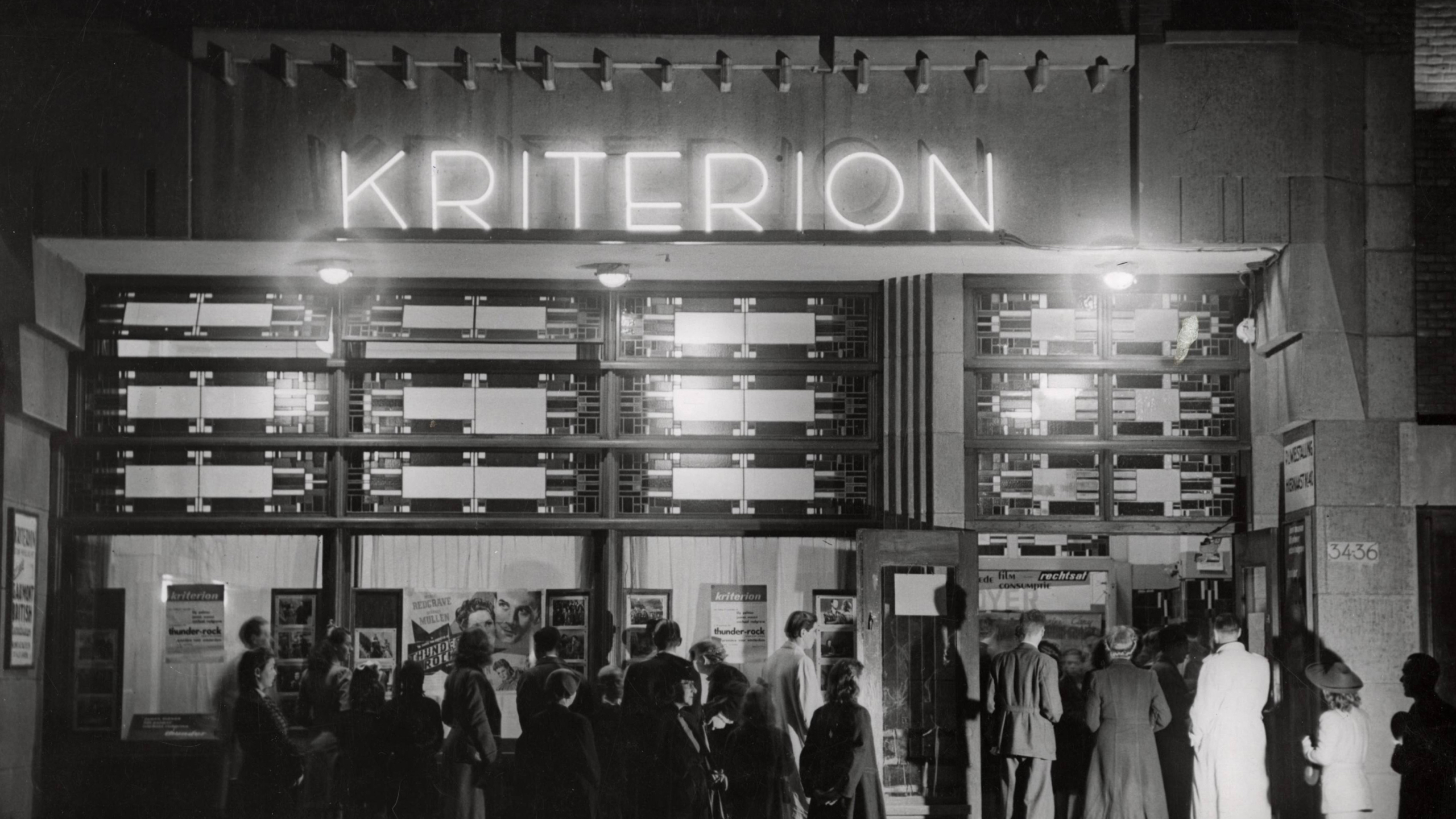 Mensen staan in de rij voor de ingang van bioscoop Kriterion, verlicht door neonletters, met posters te zien in de etalage.